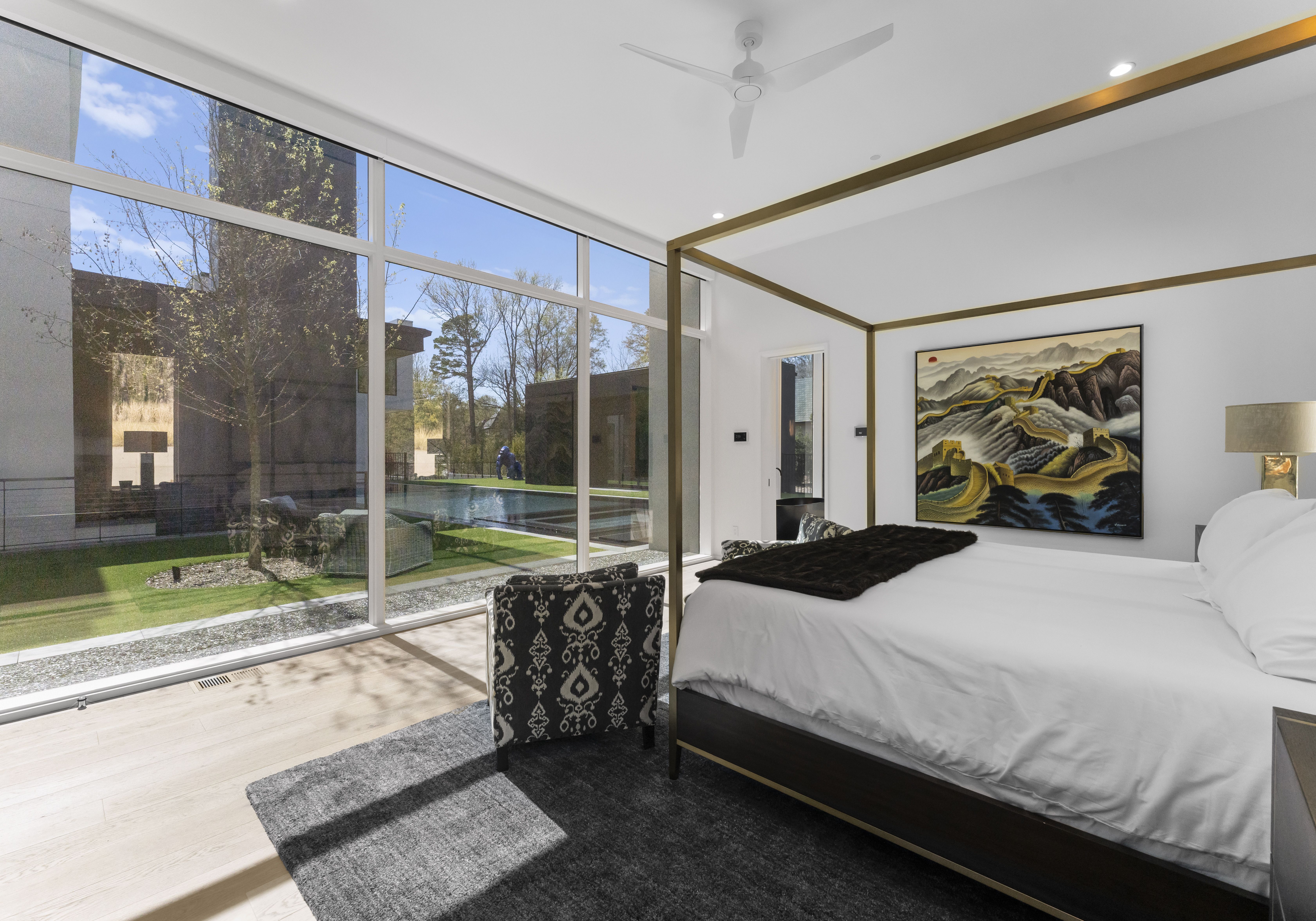 Bright modern bedroom with large floor-to-ceiling windows showing an outdoor pool and trees. Four-poster bed with white linens, dark patterned chairs, a gray rug, and a colorful mountain painting.