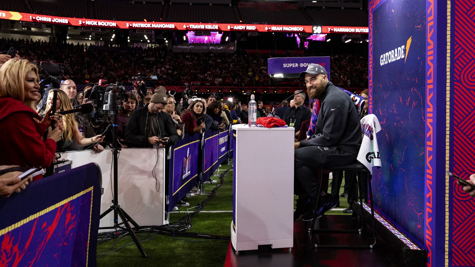 Travis Kelce sit behind a desk while a crowd of photojournalists edges in toward him beyond a barricade.