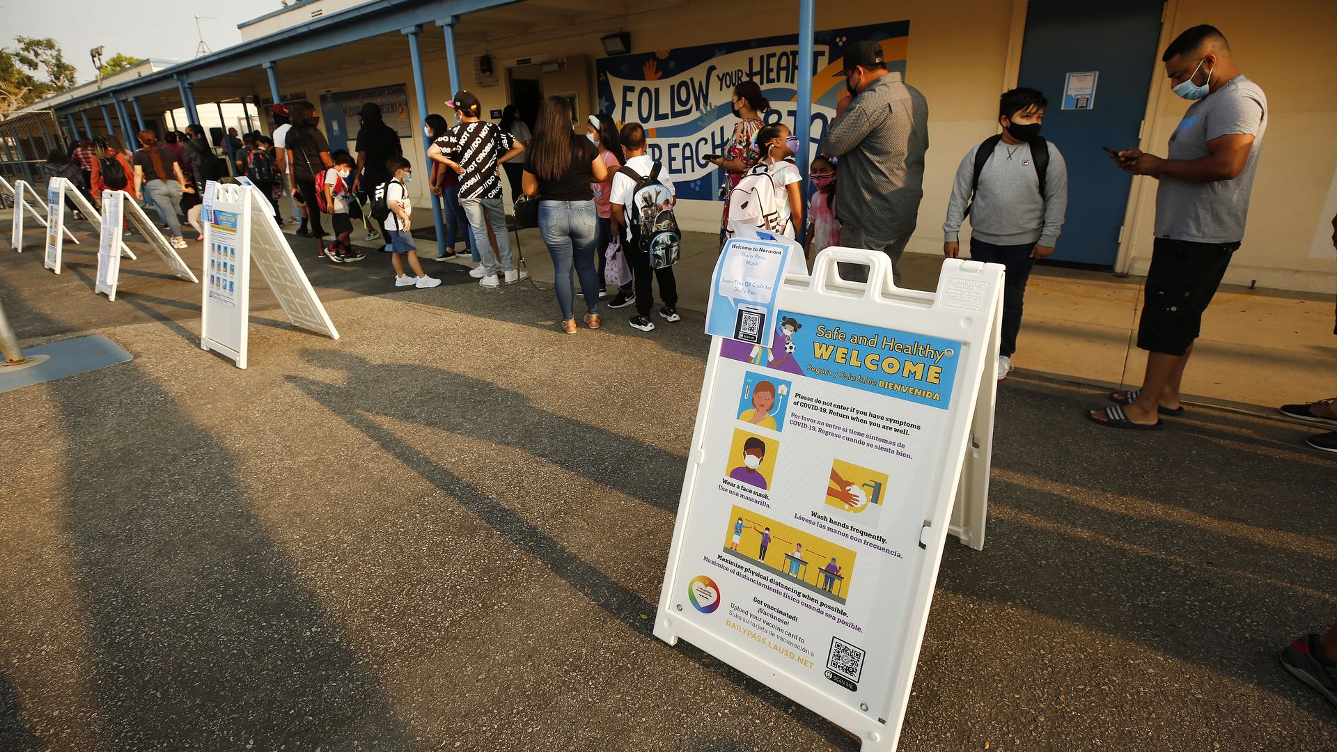 Photo of children and parents lined up outside an elementary school building, with a sign emphasizing safety and health in the foreground