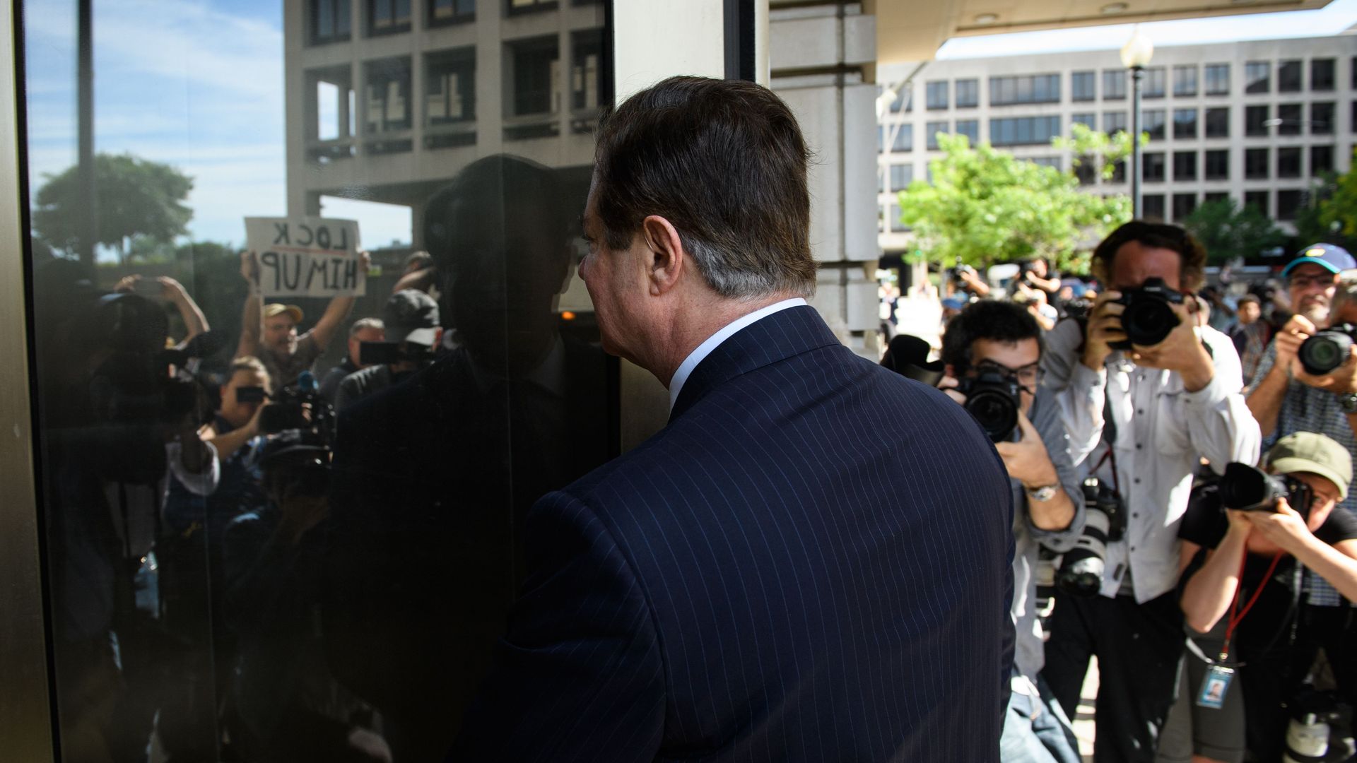 Paul Manafort walks into a building with his back toward the camera. In the revolving door reflection you can see cameras and protestors surrounding him.