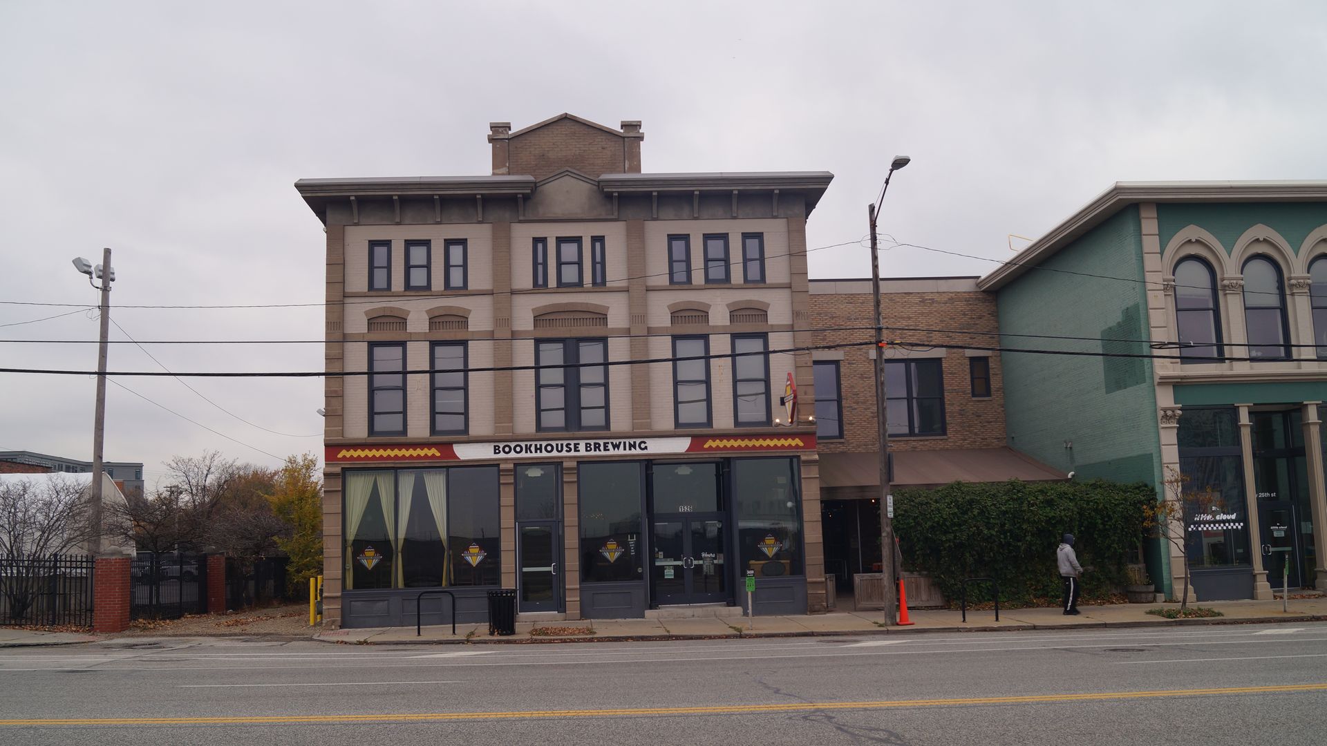 Three-story beige brick building with "Bookhouse Brewing" sign, large windows with yellow curtains, next to a green building. Overcast sky, empty street, one person walking nearby.