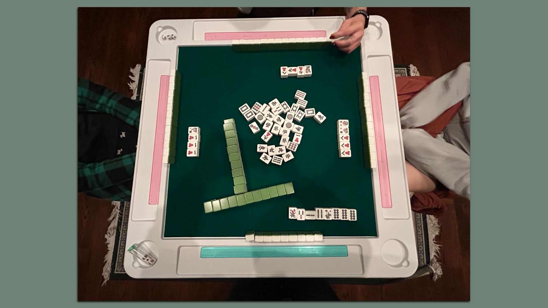 Overhead view of a mahjong game in progress on a green felt and white table with pink and blue score markers, four players visible from above with tiles arranged in front of them.