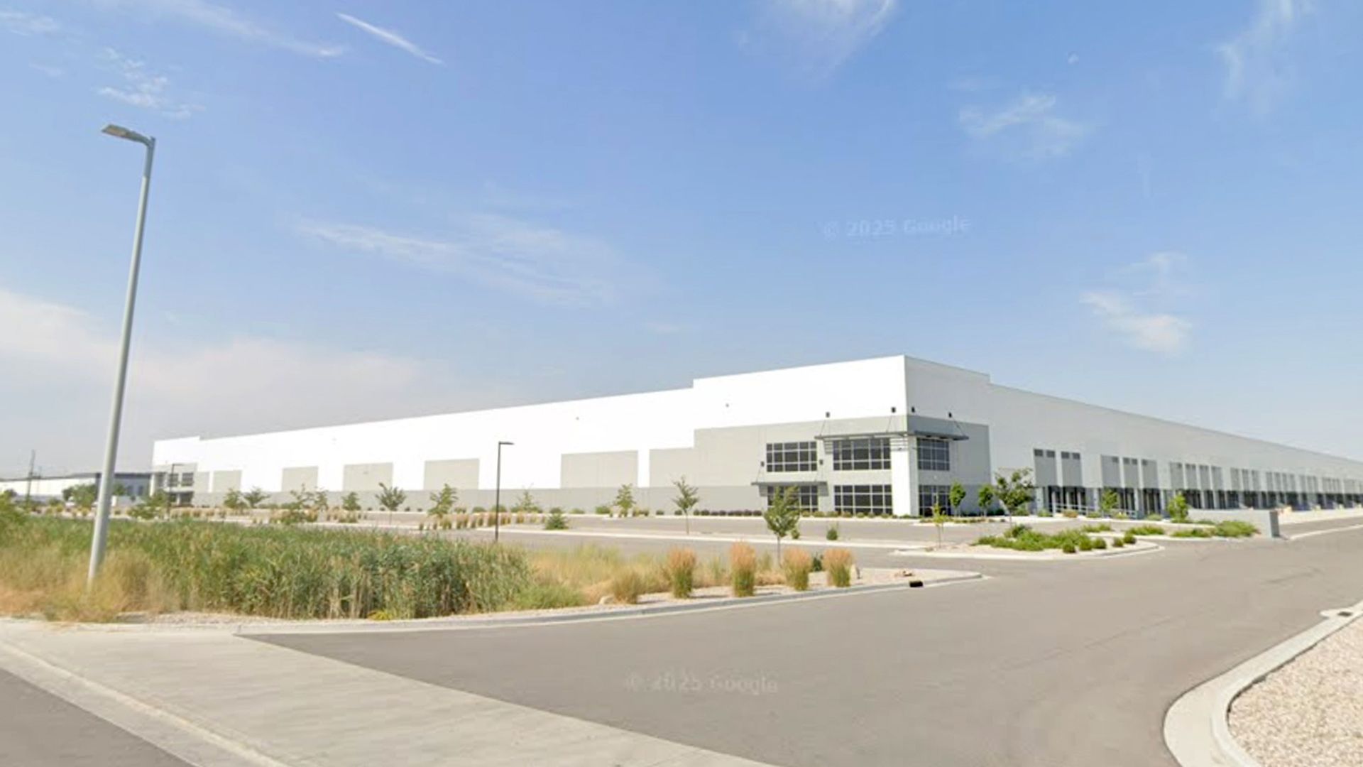 A large white industrial warehouse with multiple loading bays sits along a curved road, with small trees and grasses along the lot, a tall streetlamp, and a clear blue sky.