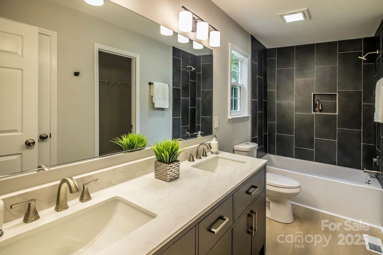 Modern bathroom with double sinks, white countertop, black cabinets, a bathtub with large dark tiles on the wall, white toilet, window, and green plant on the counter.
