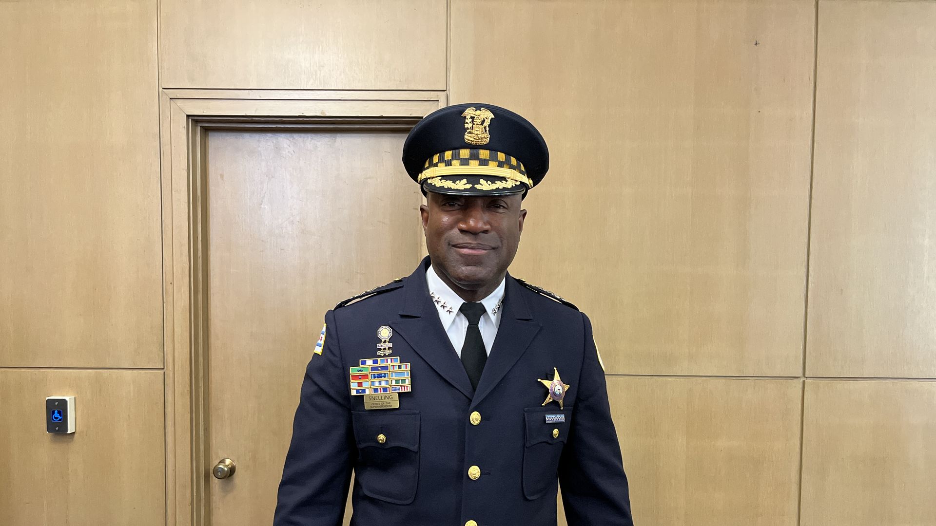 A smiling Black police officer in full dress uniform stands in a wood-paneled hallway, wearing a navy cap with gold insignia and a chest full of ribbons and medals.