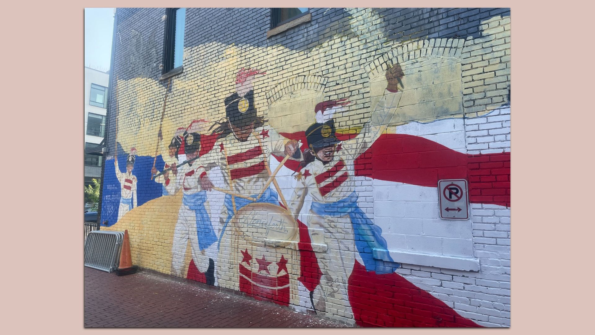 Brick-wall mural of a patriotic parade with musicians in white uniforms and red accents; a yellow drum is visible, and a No Parking sign sits on the right.