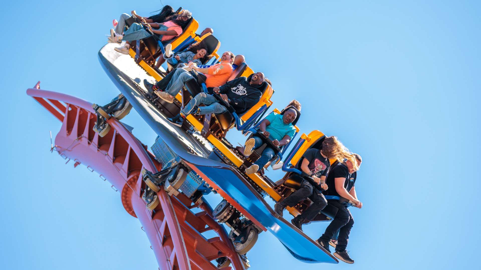 Riders in bright orange and blue seats on a twisting roller coaster, screaming with excitement against a clear blue sky; the red track and blue rails snake beneath.