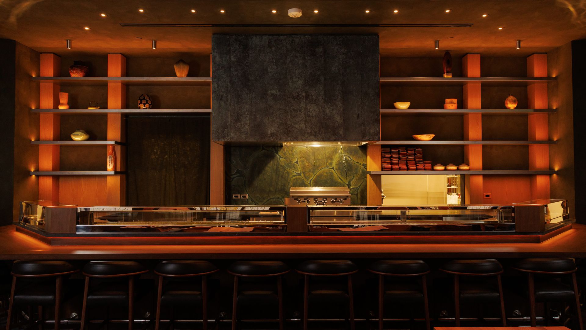 Warm, modern bar interior with amber lighting, symmetric orange shelves displaying pottery, a large central dark cabinet, and a long counter with black stools in front.