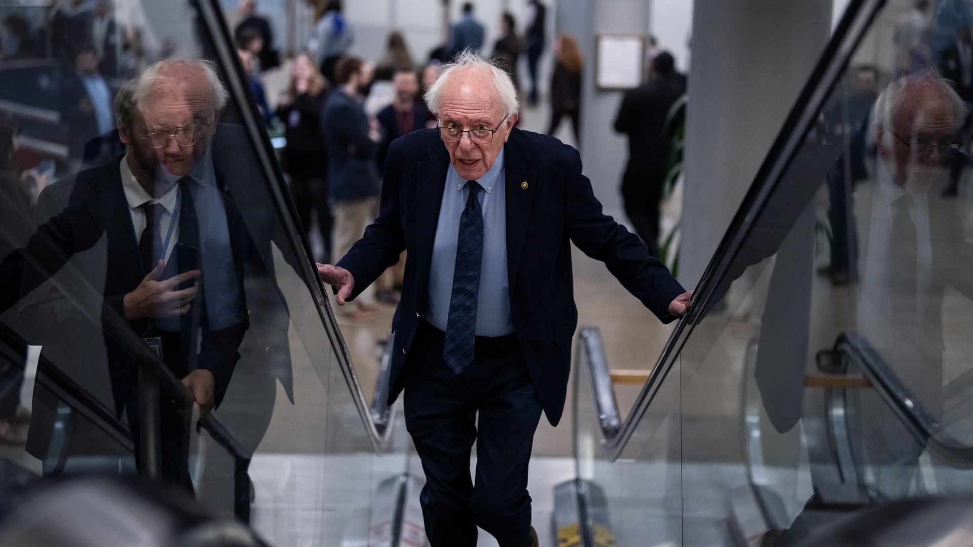 Man in a dark suit and blue tie climbing stairs indoors, with crowd of people blurred in background and reflections on glass handrails.