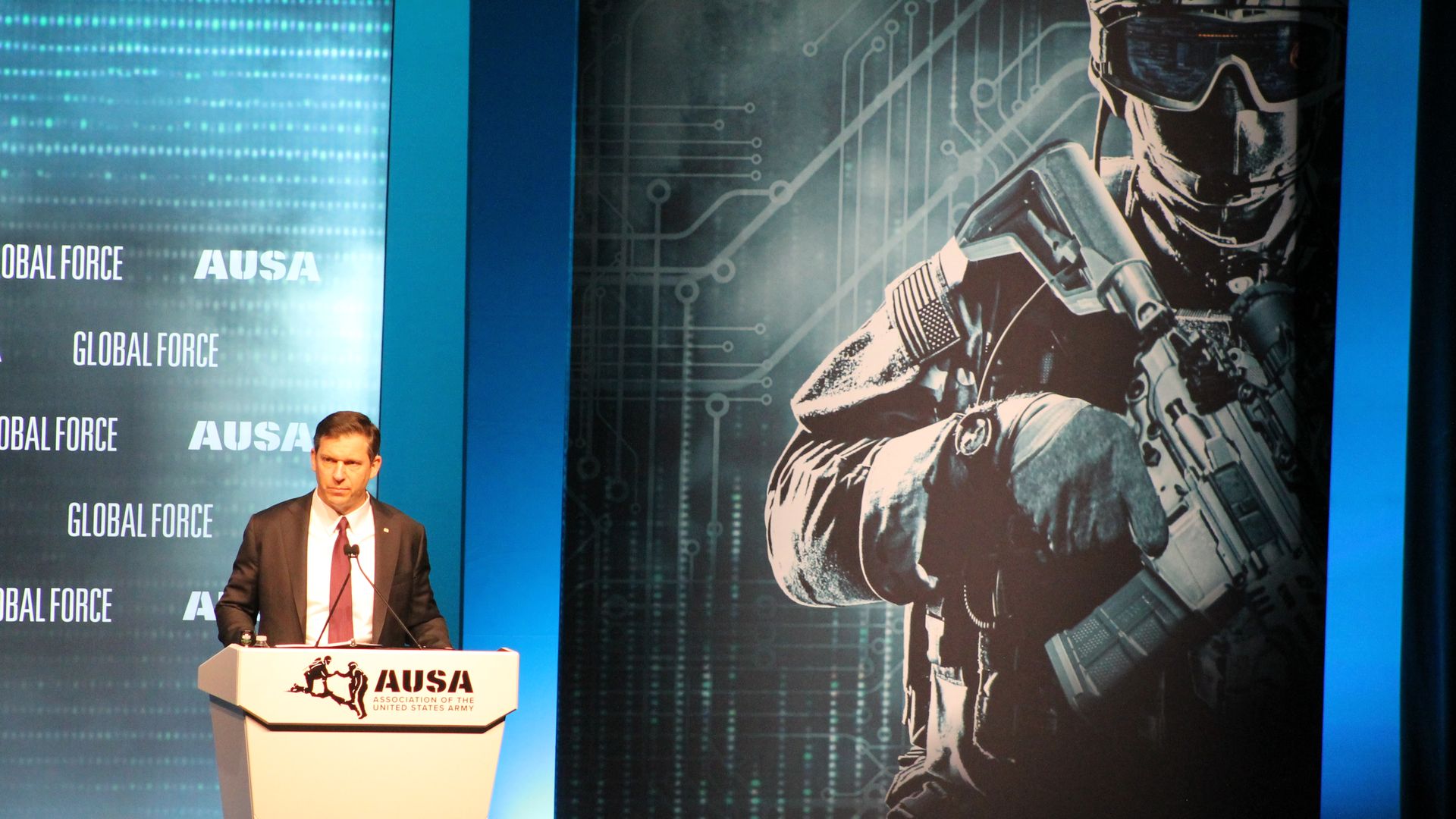 Man in a suit and red tie speaks at a podium labeled "AUSA" on a blue-lit stage; to the right, a large mural shows a soldier in tactical gear against a circuit-like backdrop and the text "GLOBAL FORCE".
