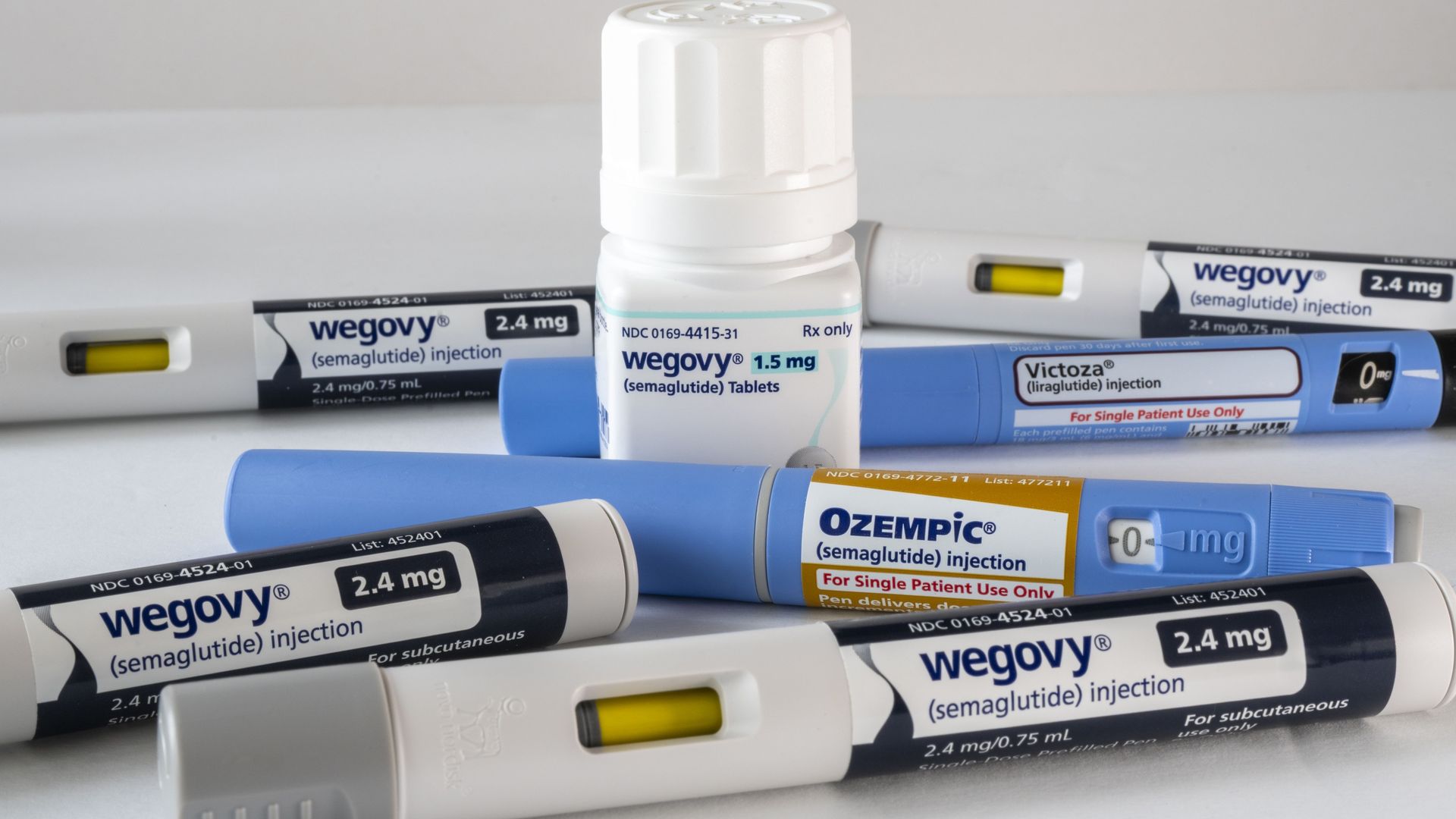 Several medication pens and a bottle: Wegovy (semaglutide) injection 2.4 mg and tablets 1.5 mg, Ozempic (semaglutide) injection, and Victoza (liraglutide) injection on a white surface.