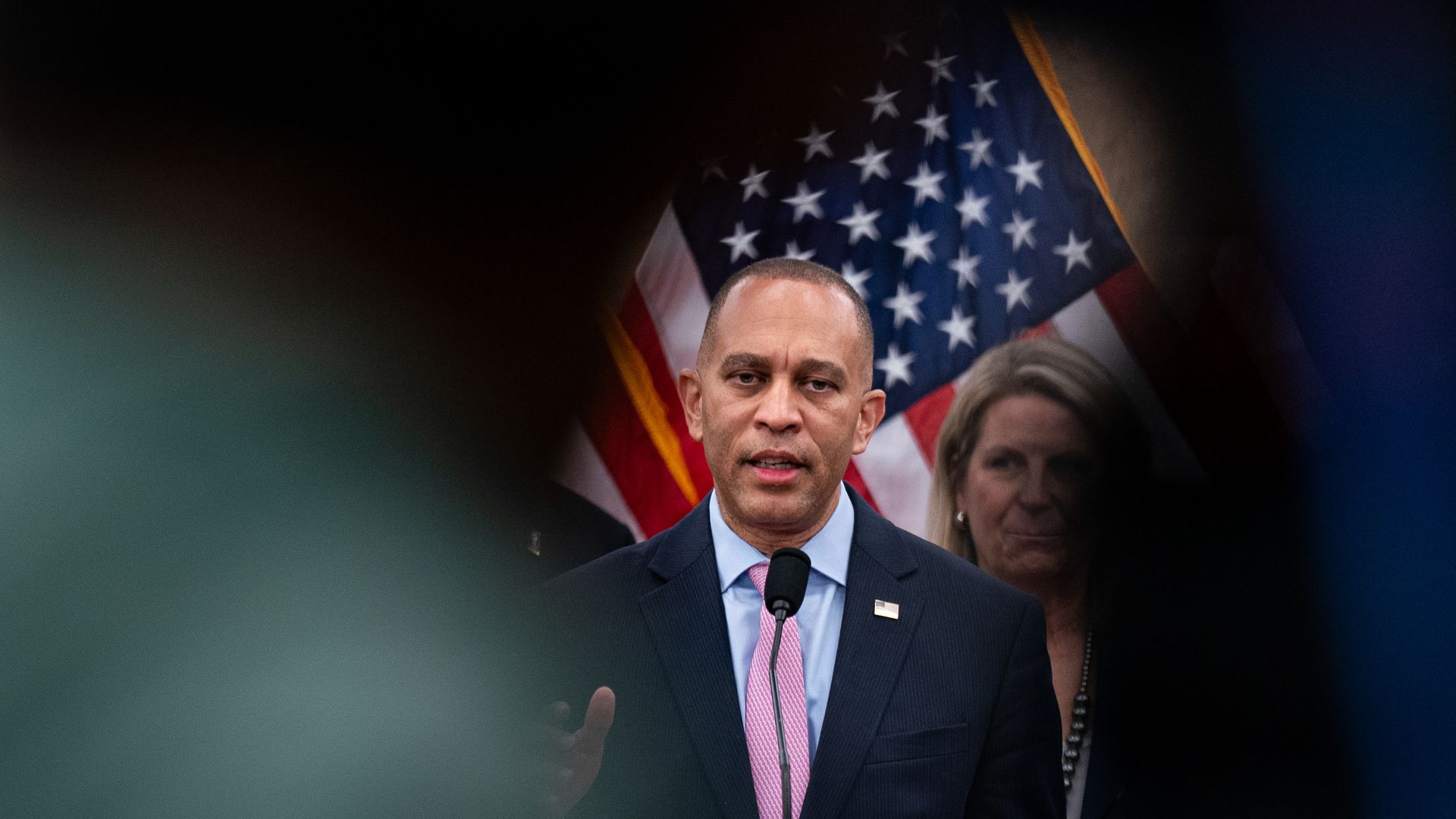 Hakeem Jeffries, wearing a blue suit, speaking into a microphone in front of an American flag.