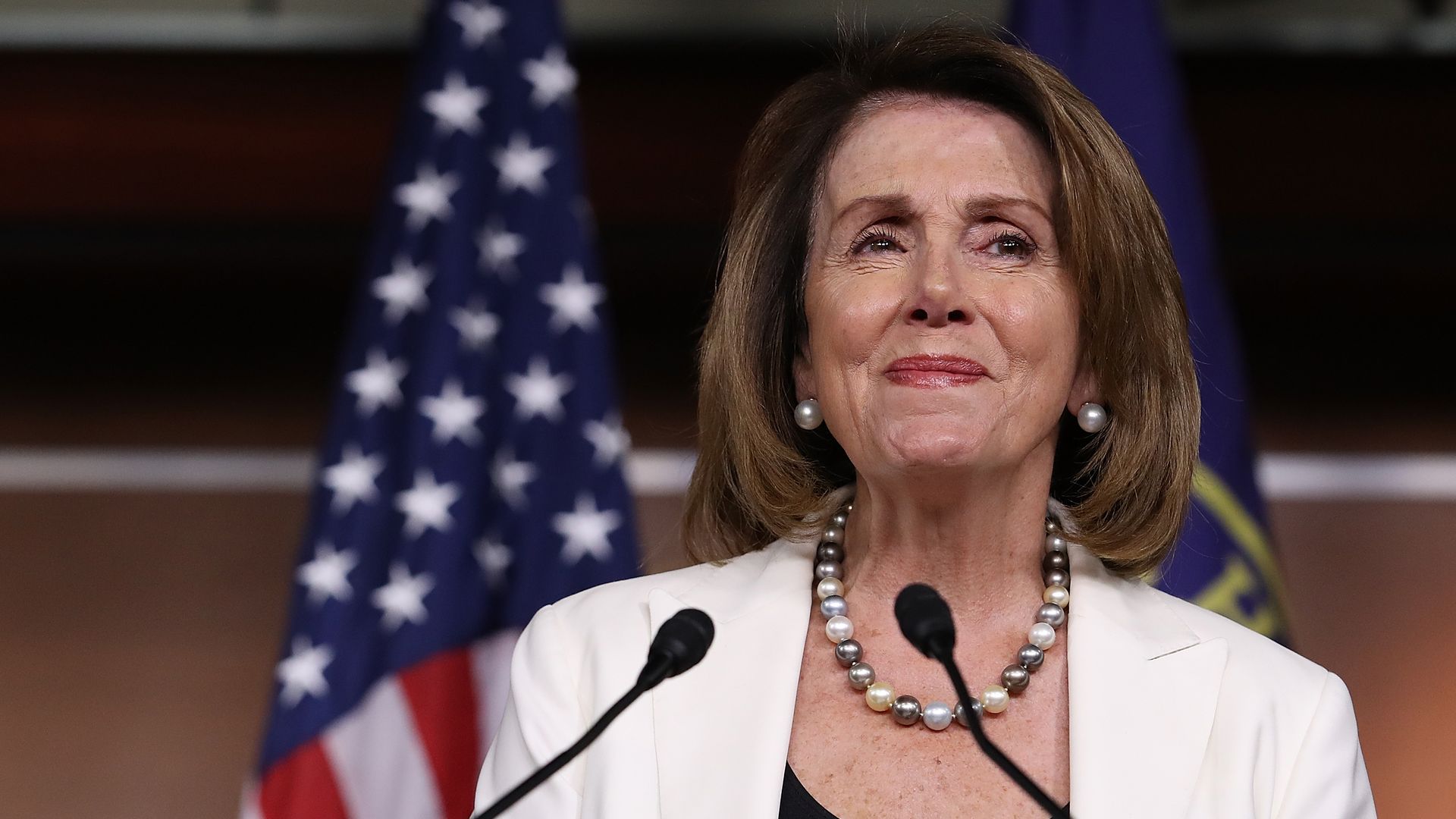 Nancy Pelosi in a white suit standing at a podium with a proud smile on her face.