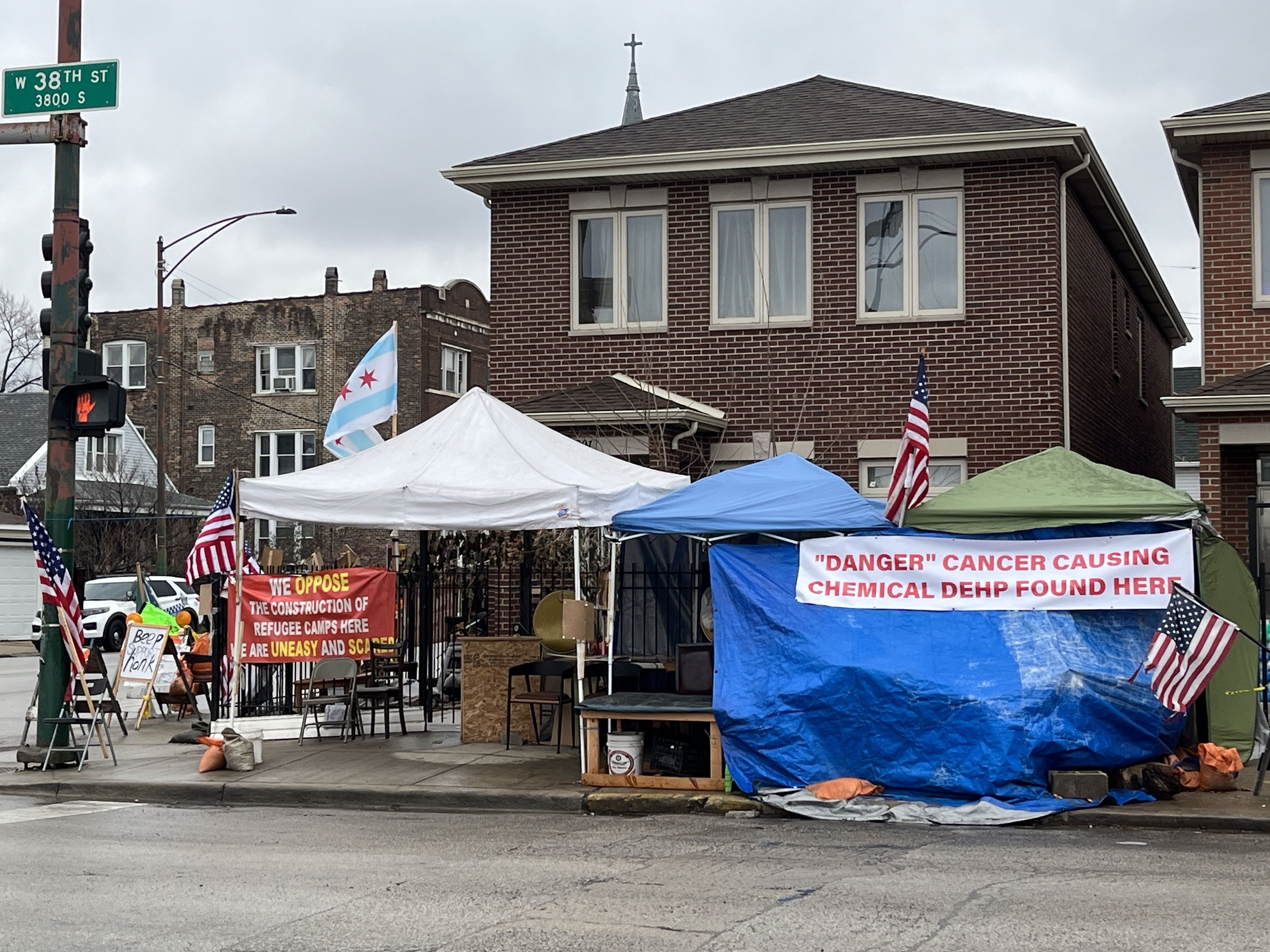 Two small tents stand on the sidewalk with protest signs objecting to environmental concerns at the Brighton Park migrant camp.