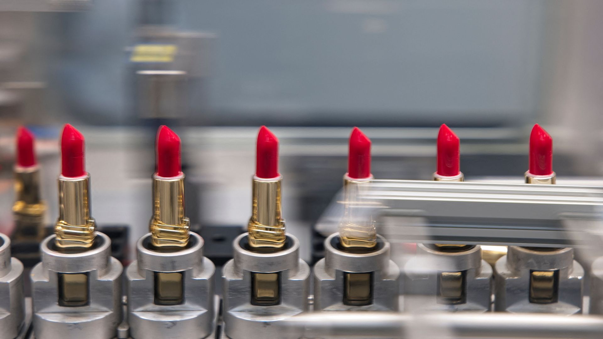 Row of bright red lipsticks with gold bases on a production line in a factory setting with a gray background.