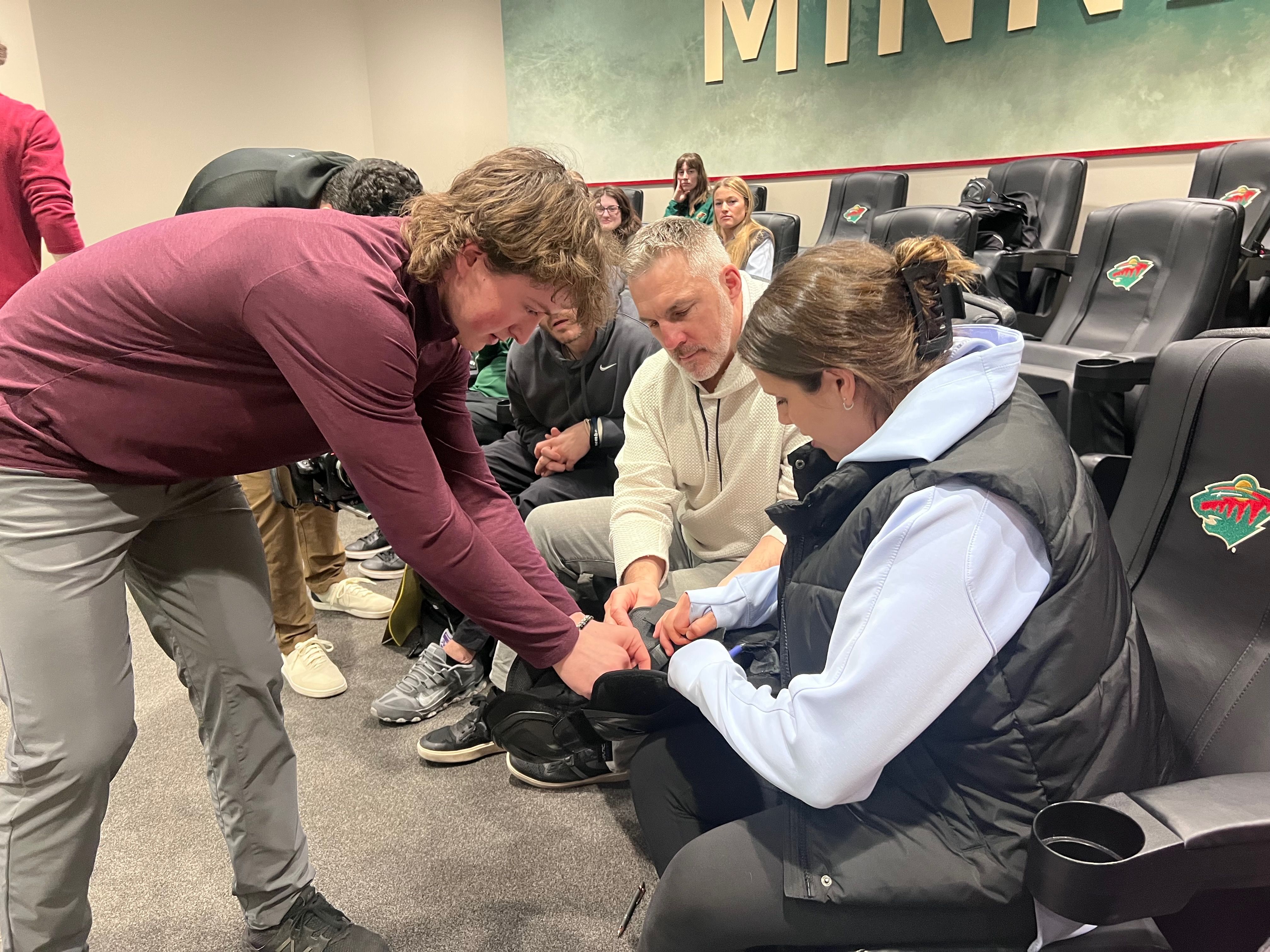 A group of adults seated in a room with Minnesota Wild logos on chair backs. A man in a beige sweater and a woman in a blue hoodie vest assist with a shoe while another person in maroon leans in.