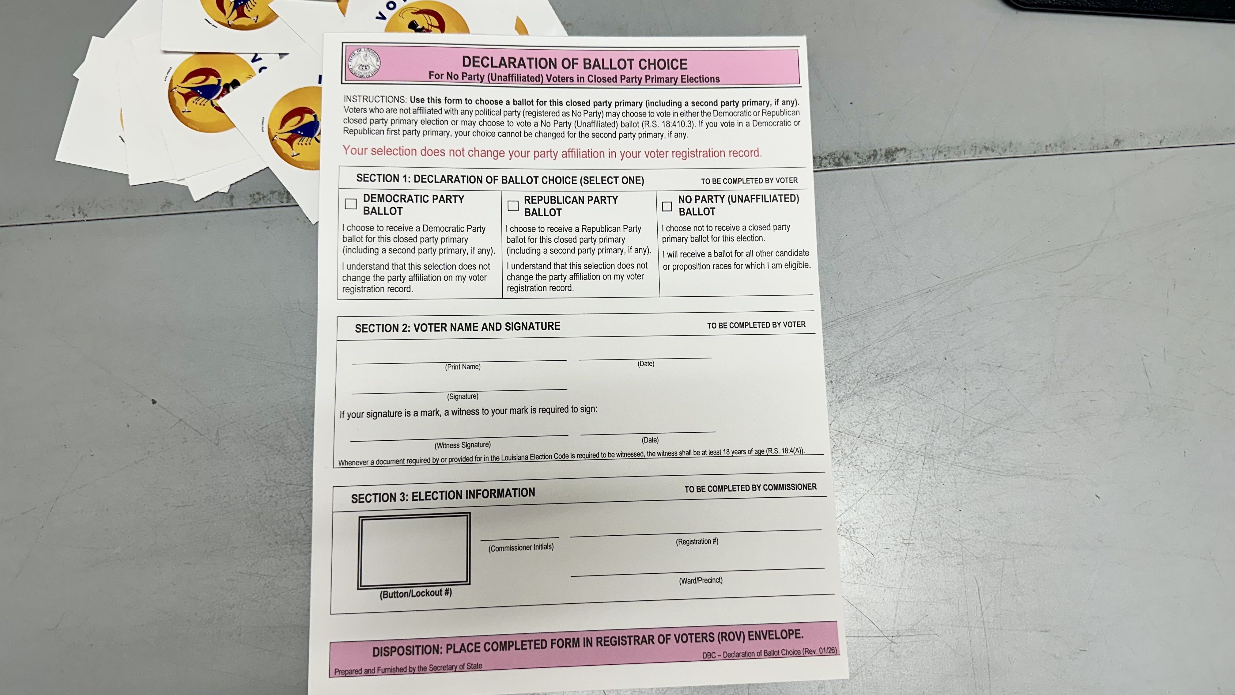 Close-up of a pink-header "DECLARATION OF BALLOT CHOICE" ballot form on a gray desk, showing checkboxes for Democratic, Republican, No Party; stacked stickers with orange-yellow logos to the left.
