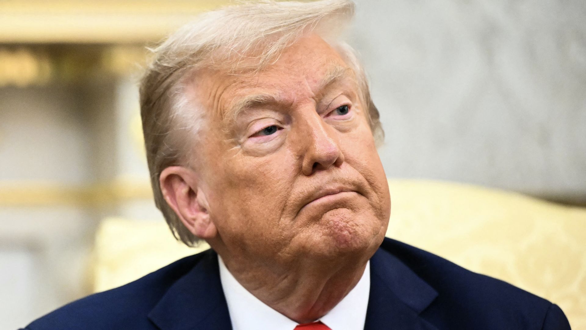Close-up of a man in a navy suit, white shirt, and red tie with an American flag pin, looking upward with a serious expression. Background is softly blurred, indoors.