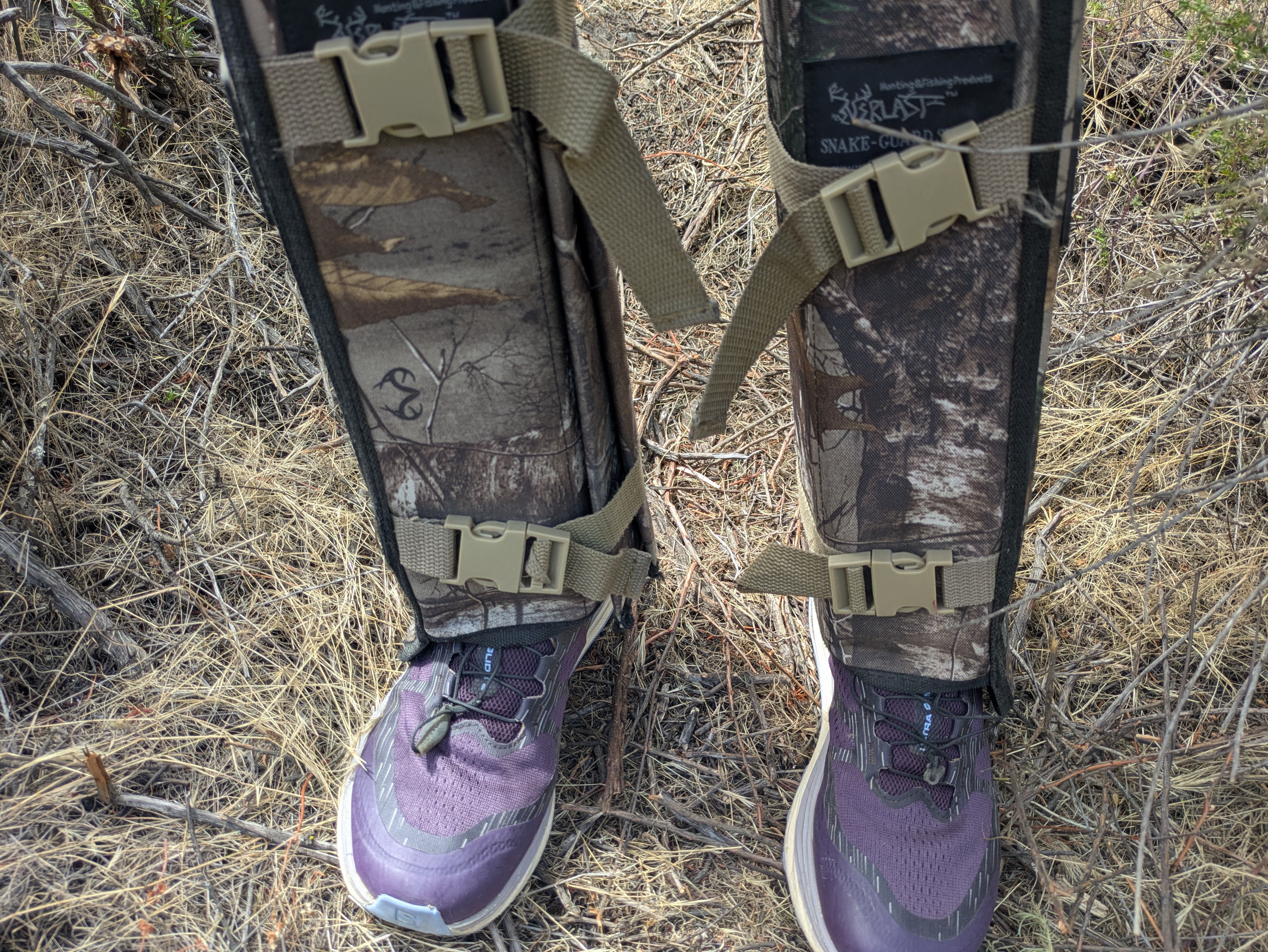 Close-up of two legs wearing camo gaiters with tan buckles over purple sneakers, standing in dry grass and twigs.