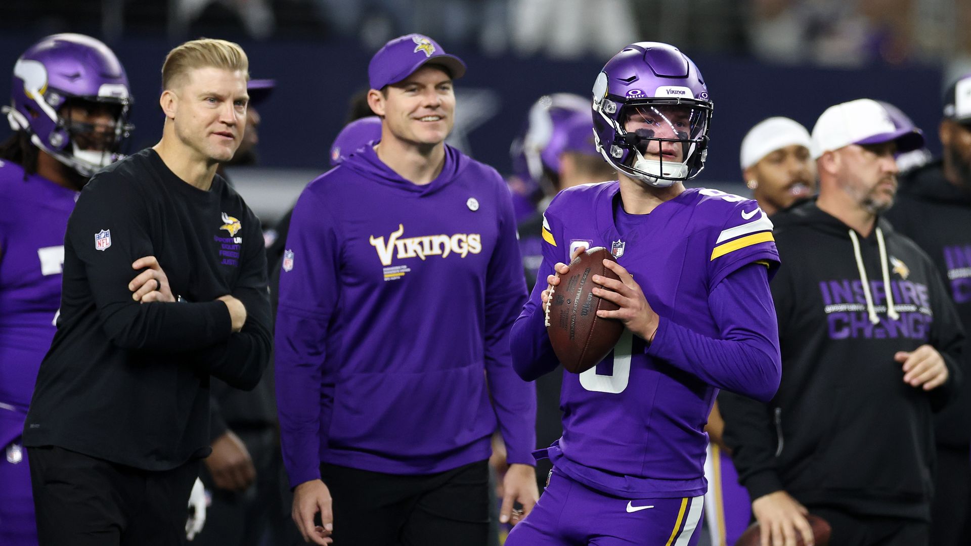 JJ McCarthy is wearing a purple uniform and getting ready to throw a football while Josh McCown and Kevin O'Connell watch from behind him. 