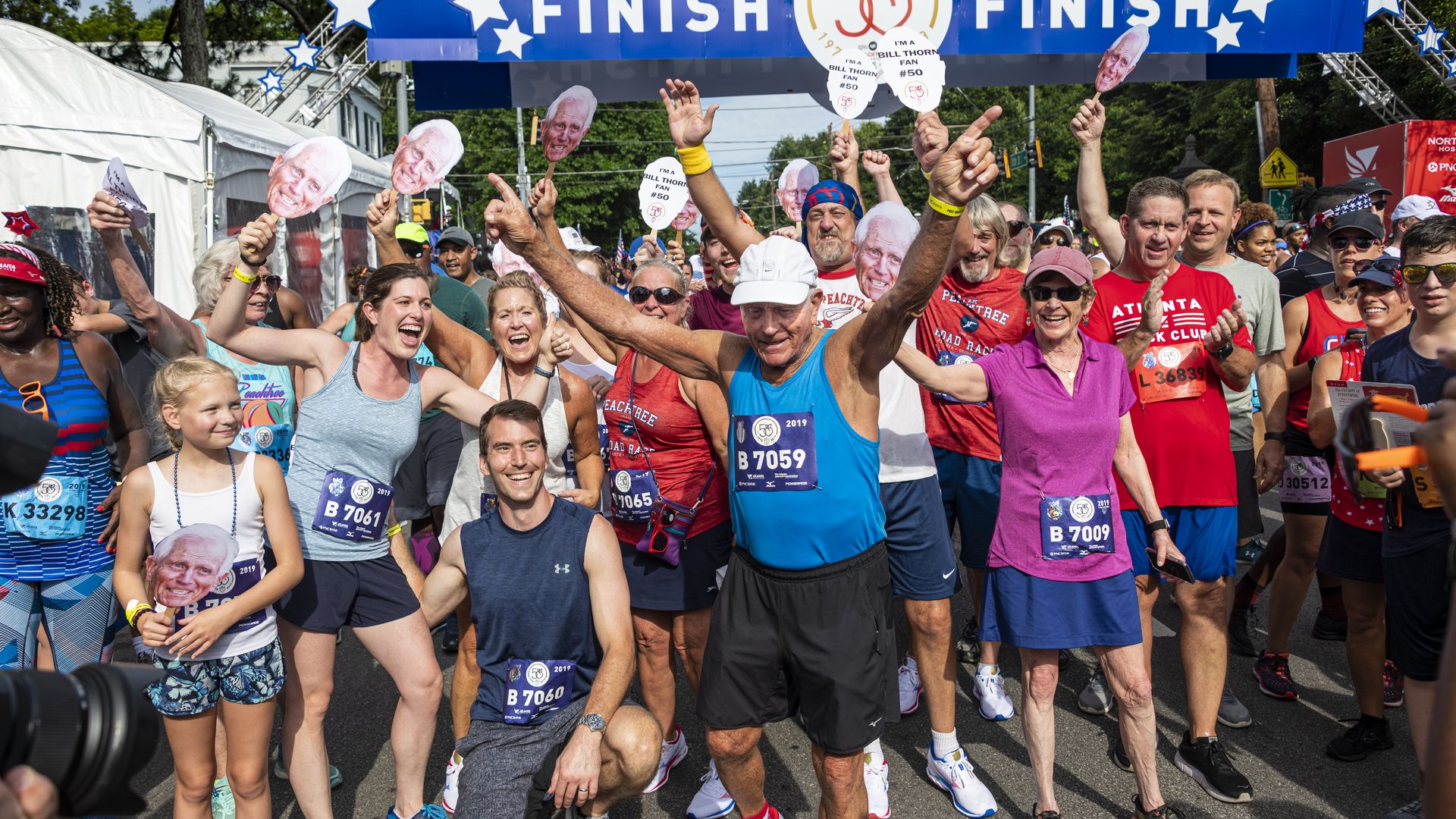 Bill Thorn, a 92-year-old man wearing a tank top and running shorts, celebrates finishing a race with his arms outstretched while people behind him cheer
