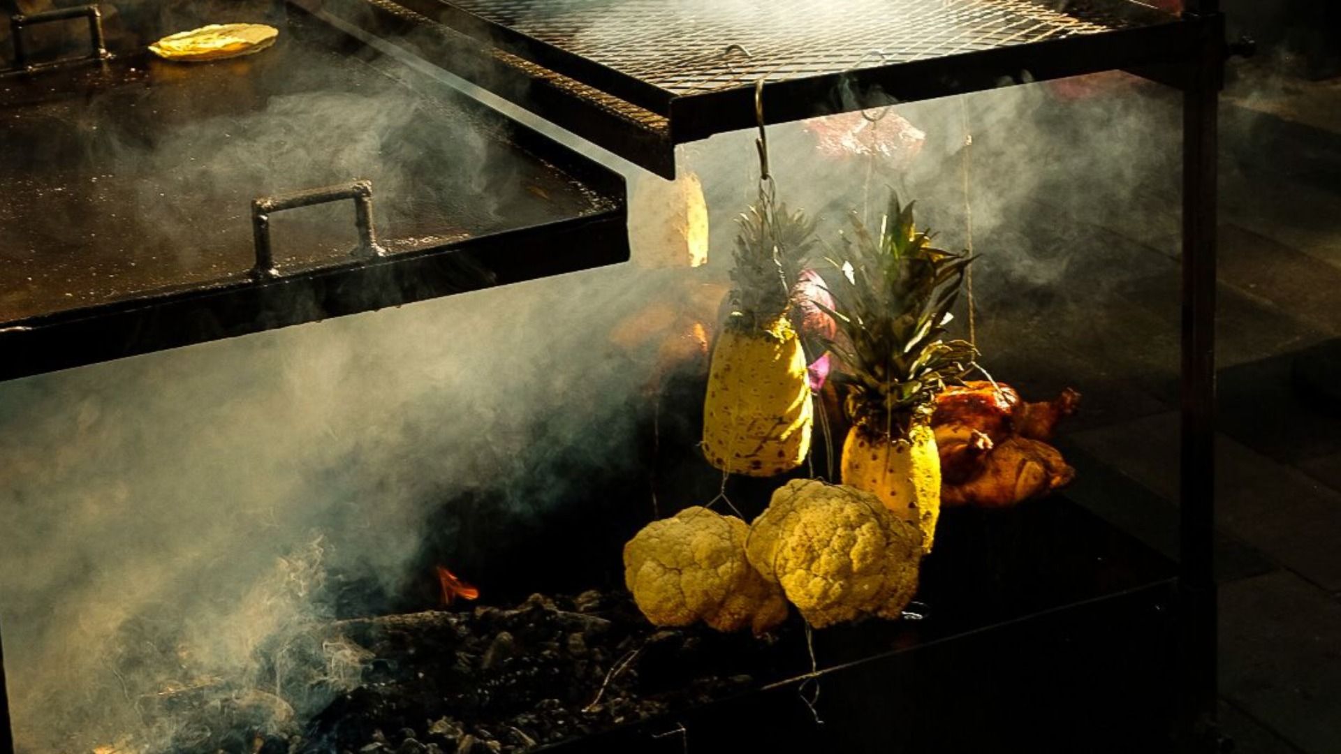 Smoky grill scene: a black metal barbecue with a grate overhead; pineapples and cauliflowers hang from hooks above glowing charcoal.