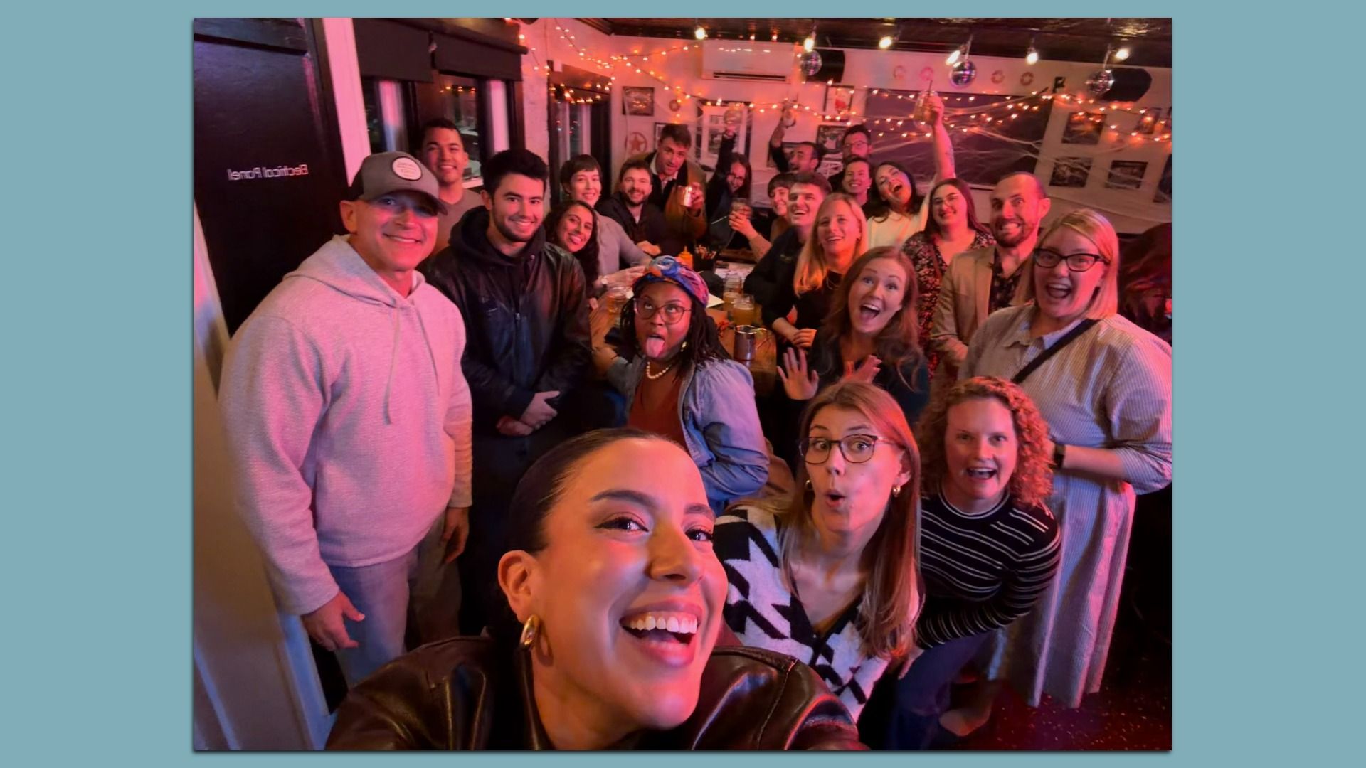 Large group of friends at a cozy party posing for a selfie around a long table under warm string lights, with colorful decorations and drinks, everyone smiling and cheering.