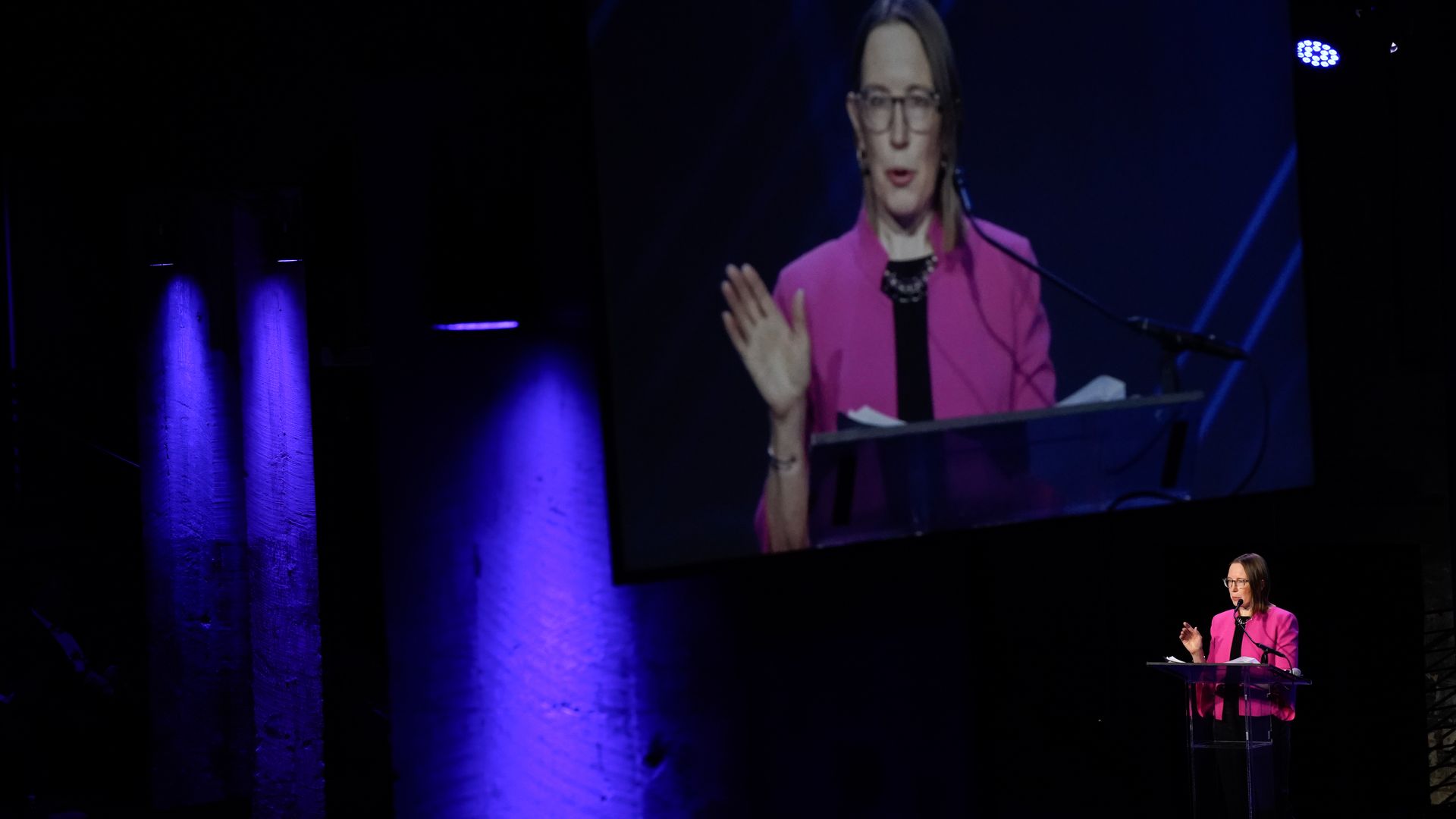Woman in pink blazer speaking at a clear podium on stage with blue spotlights and her image projected on a large screen behind her.