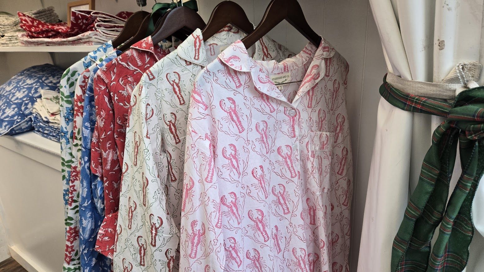 Five button-up pajamas with collars hang on a rack, featuring lobster and floral prints in red, blue, white, and green shades, next to a white curtain tied with a green plaid ribbon.
