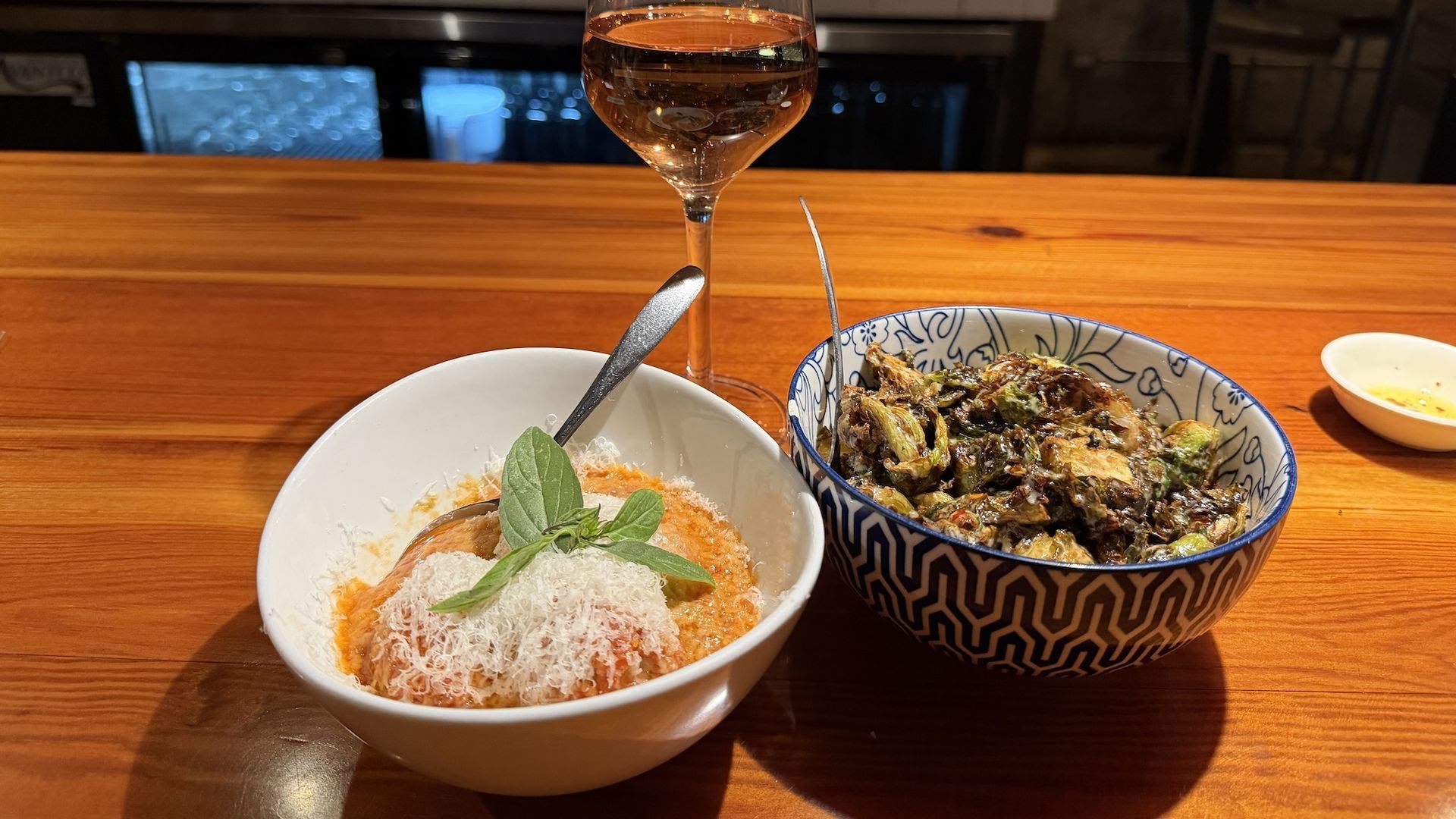 On a warm wooden table, white bowl with orange sauce, shaved cheese, and basil; blue-patterned bowl with roasted vegetables. A tall rosé glass sits behind, with a small side dish to the right.