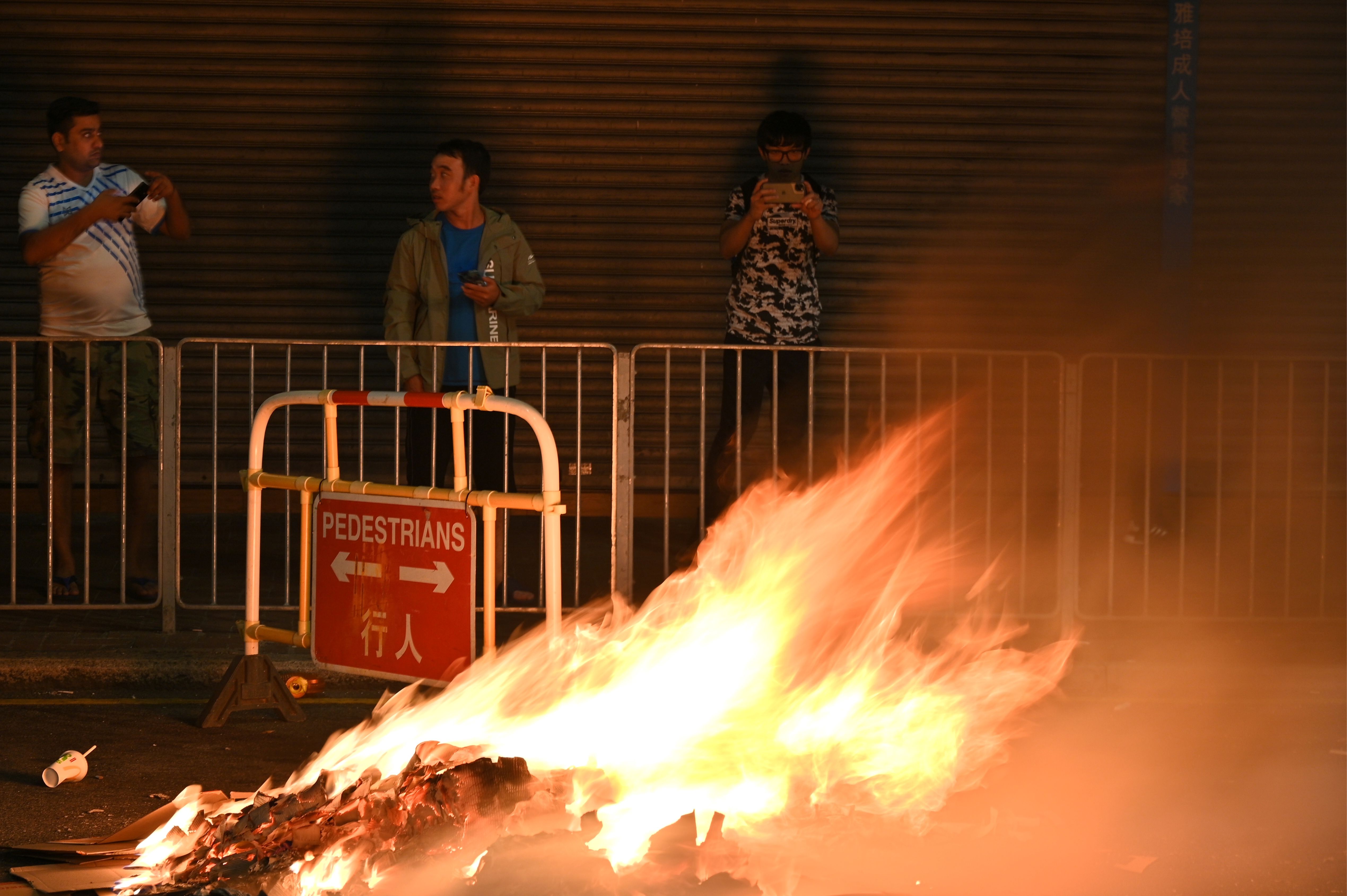 Bystanders record footage with their mobile phones as a barricade set up by protesters burns.