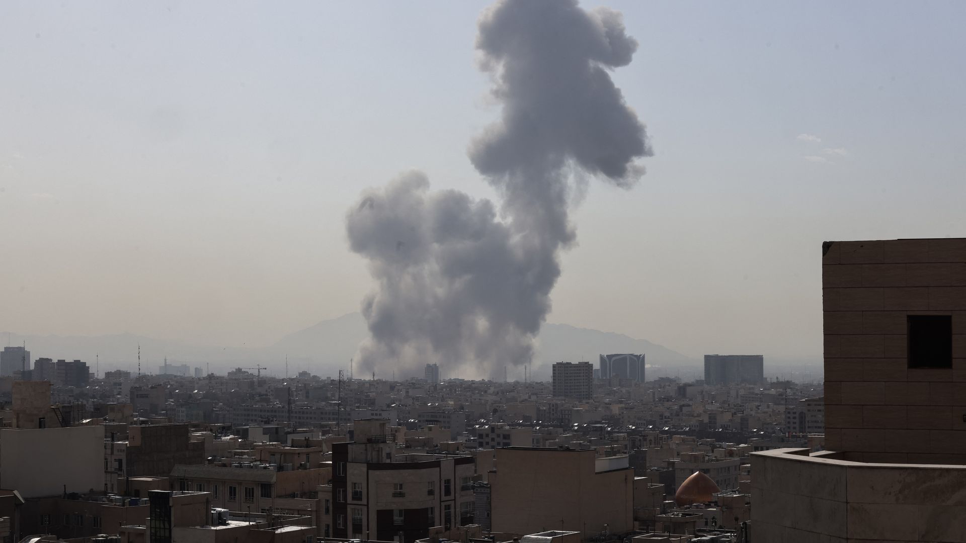 Large plumes of gray smoke rise above a cityscape with multiple buildings under a hazy sky, suggesting an explosion or fire in the distance.