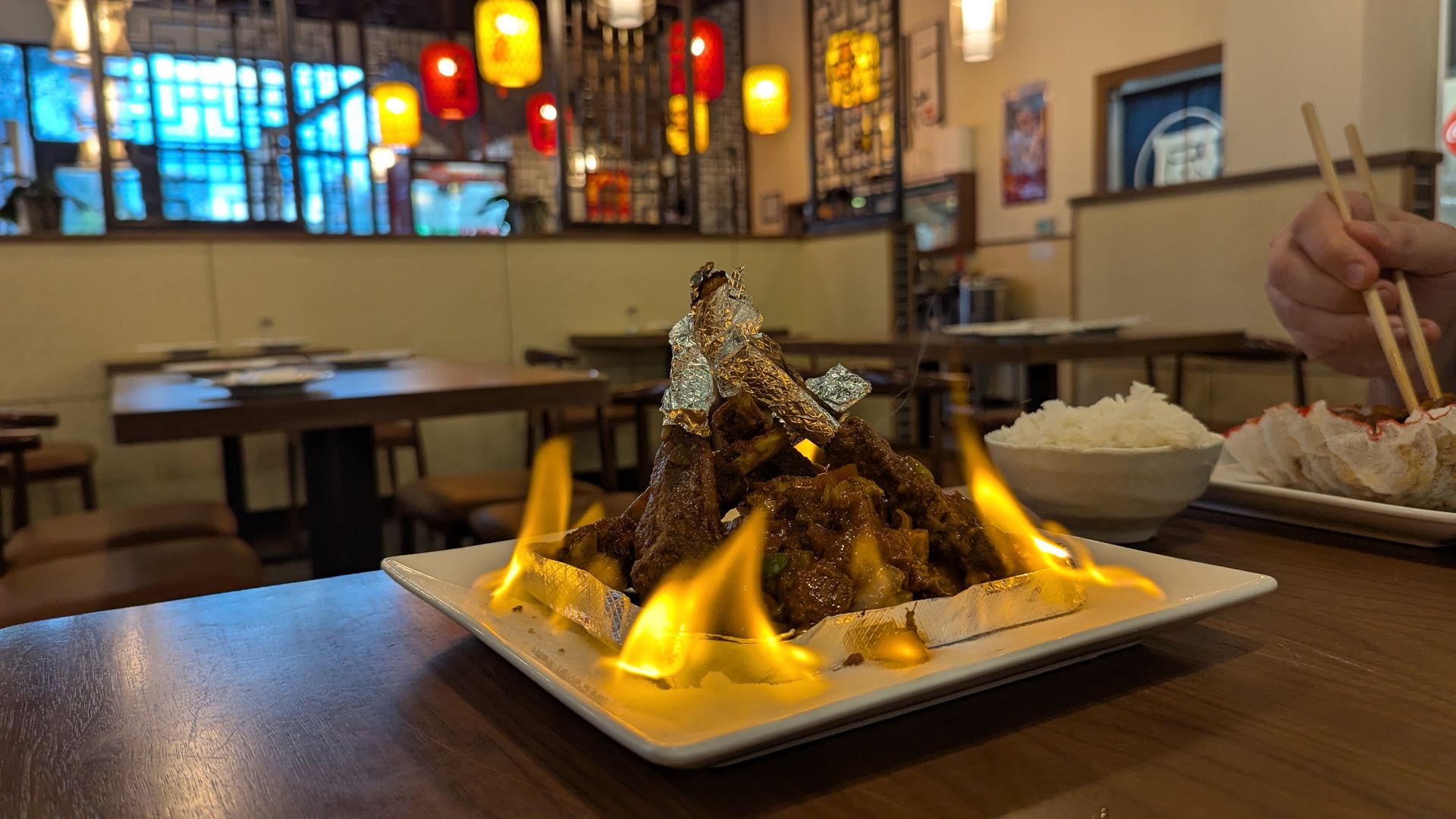 Flaming grilled meat wrapped in foil on a white plate in an Asian restaurant with glowing red and yellow lanterns. Hand holding chopsticks near a bowl of rice and dumplings.