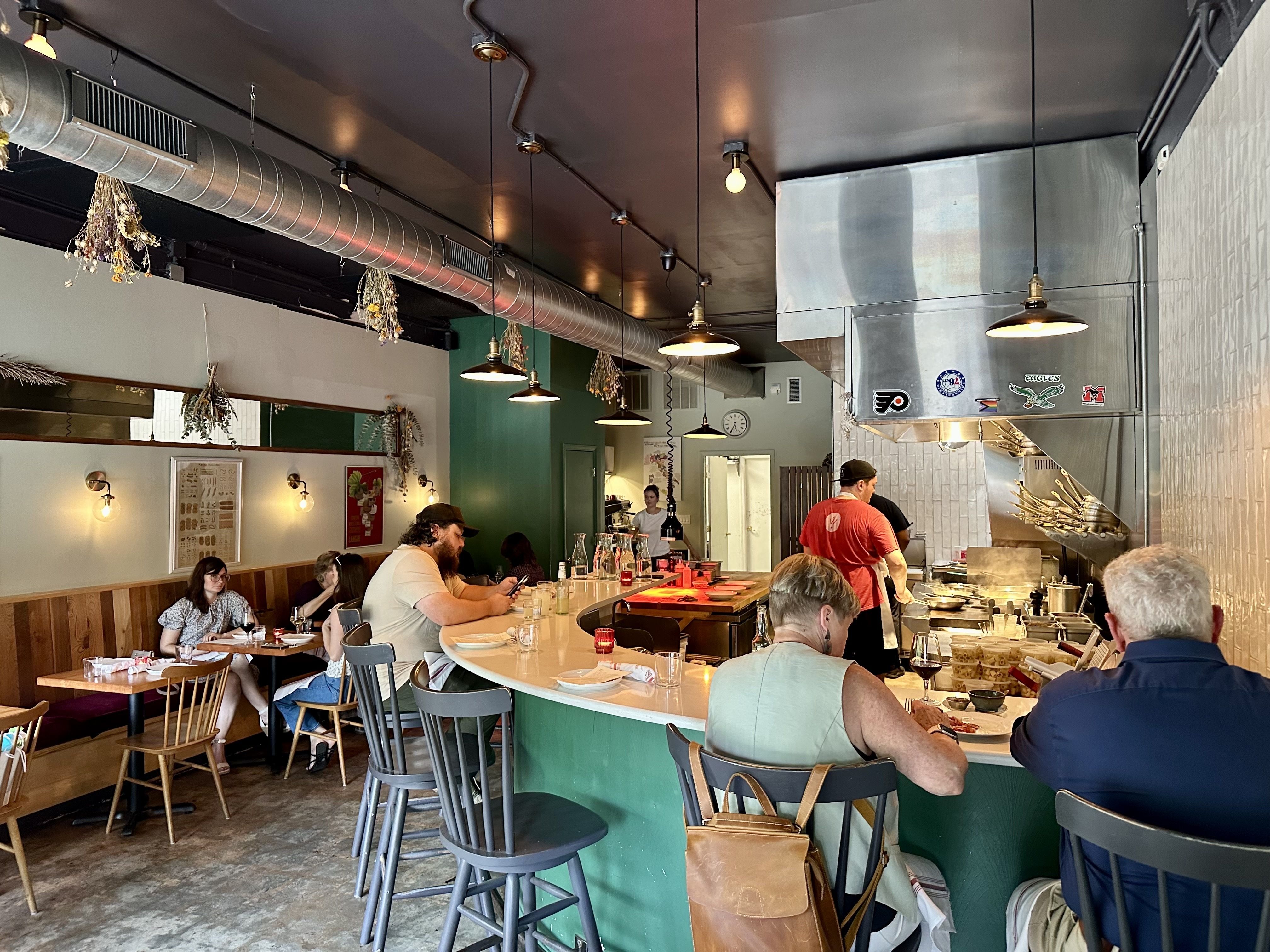 Inside a casual eatery with a long curved white counter, metal ductwork overhead, hanging bulbs, dried herb bundles, and diners seated at tables; a stainless kitchen area is visible on the right.