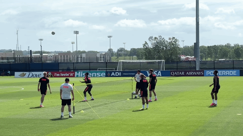 Soccer practice on a bright field with several players in dark jerseys near a goal, a ball midair, cones and training poles, and sponsor banners along the fence.