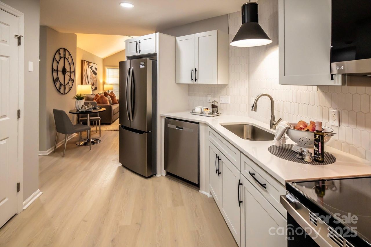 Bright kitchen with white cabinets, stainless steel fridge and dishwasher, white hexagonal tile backsplash, wooden floor, black pendant light, and a small dining area with two gray chairs and a round table.