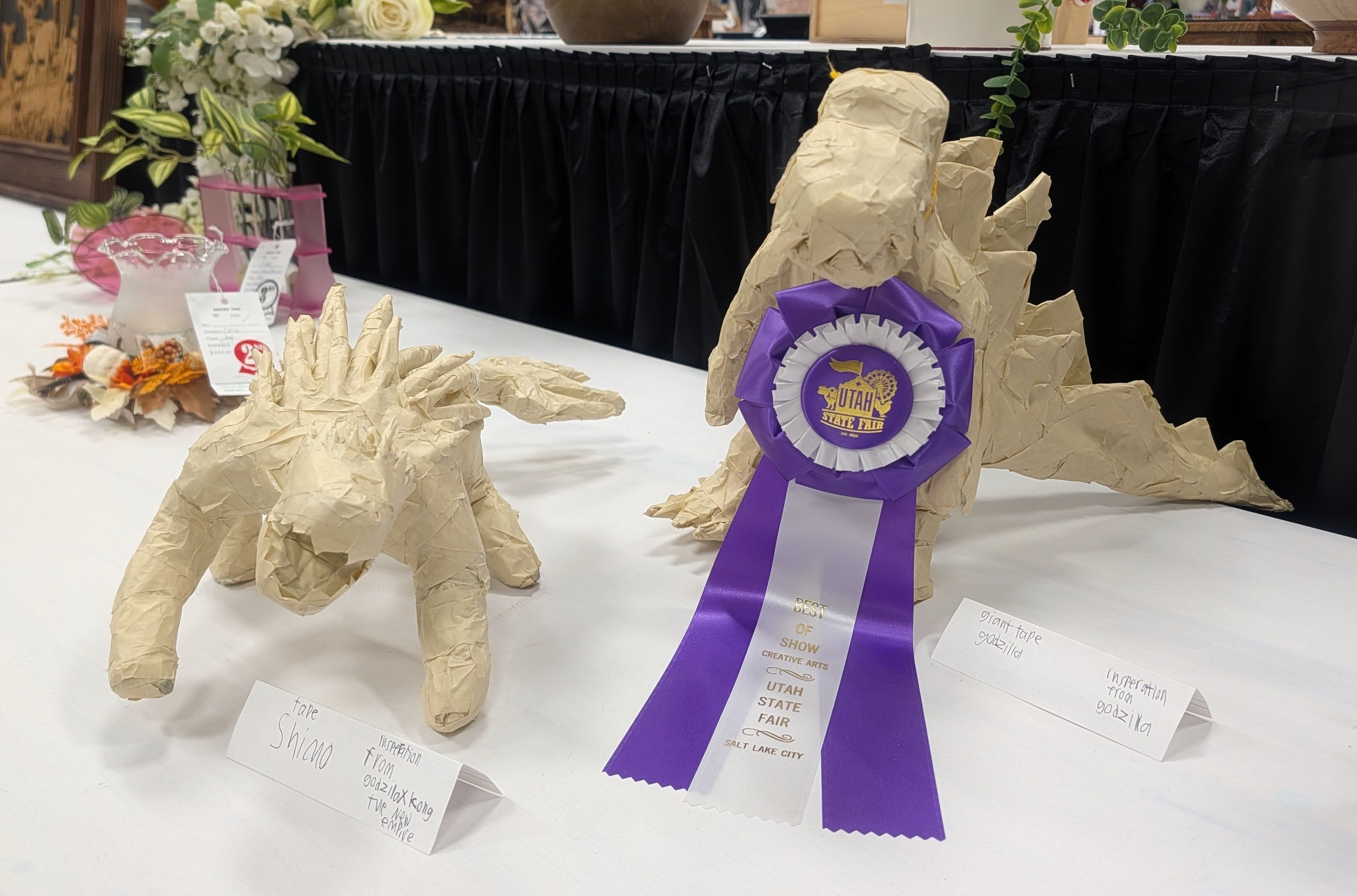 Beige paper-mâché animal sculptures on a white display table, including a large figure with a purple "Best of Show" ribbon. Floral arrangements and a black draped backdrop in the background.