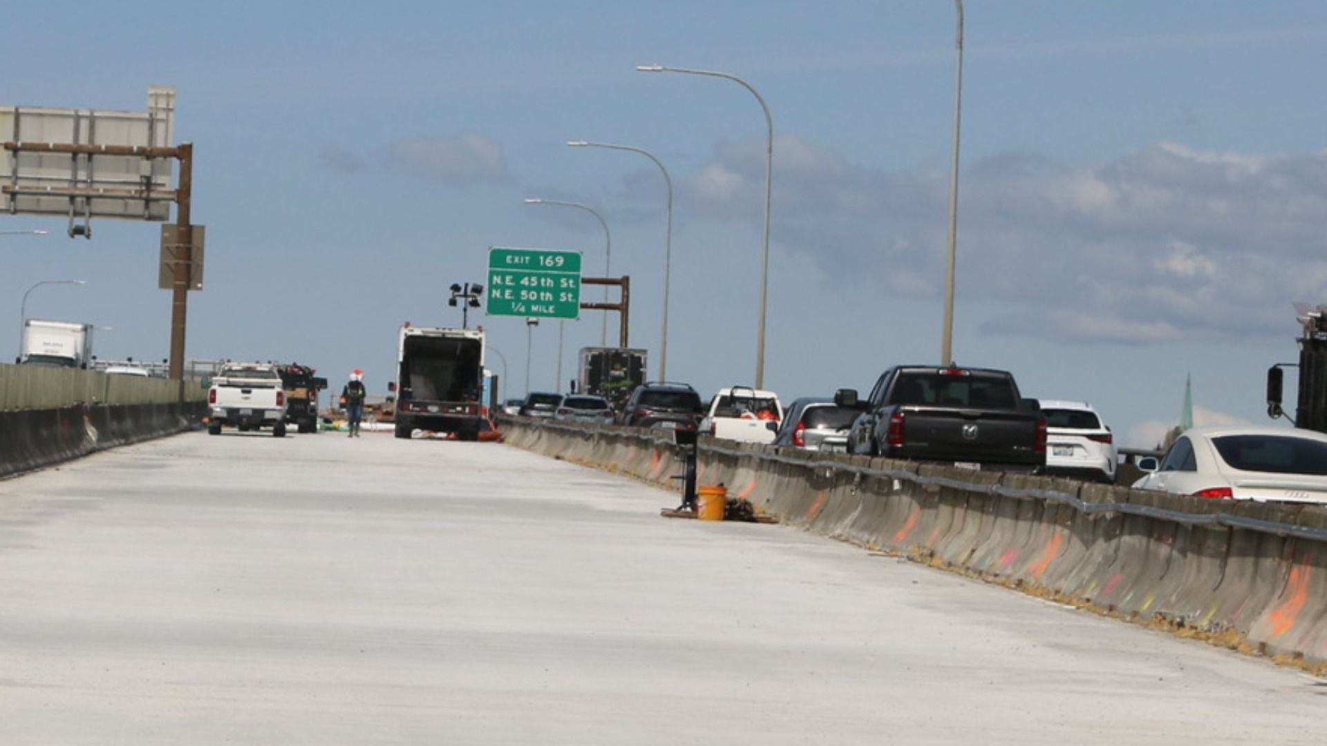 Daytime highway construction with concrete barriers, orange cones, and workers; traffic in right lanes under a green EXIT 169 sign against a blue sky.