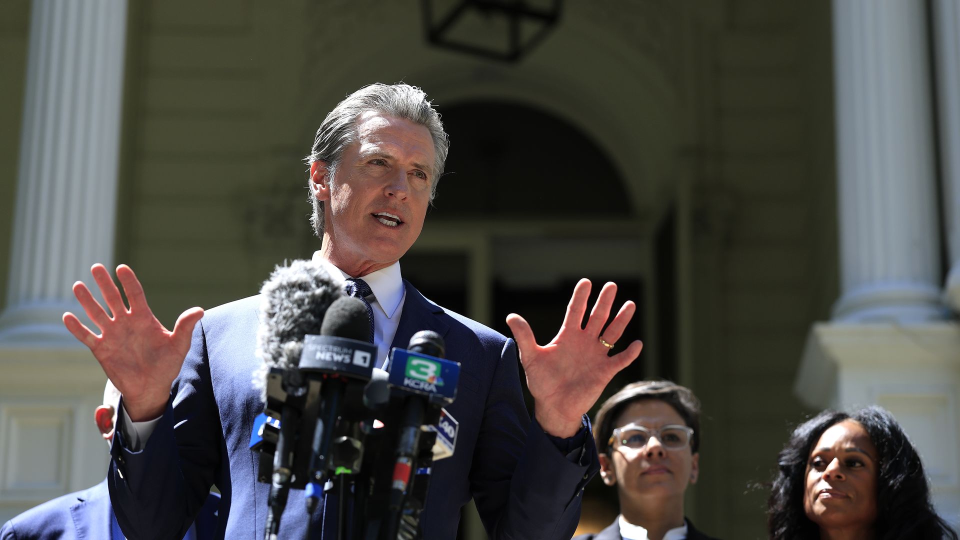 A man in a dark blue suit speaks passionately at a podium with multiple microphones; two women stand behind him, one wearing glasses and a black blazer, the other with long black hair.