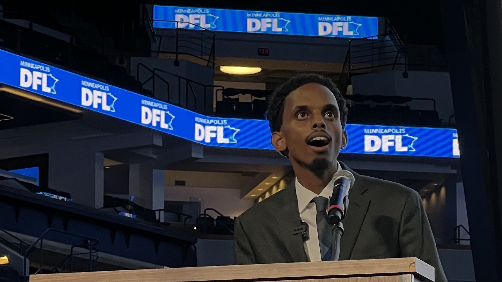 A man in a dark suit and tie stands behind a wooden podium speaking into a microphone, with illuminated blue panels in the background displaying "MINNEAPOLIS DFL" text and a state outline.