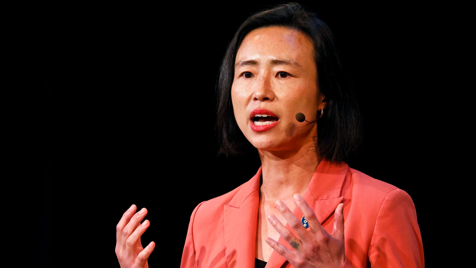 Close-up of a woman with black hair, wearing a coral blazer and headset microphone, speaking at a podium with raised hands on a dark background.