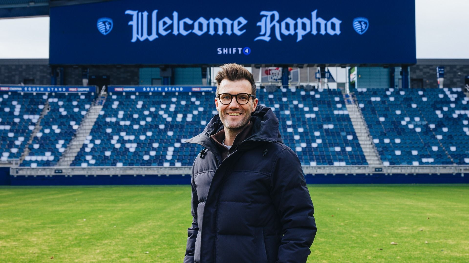 Man wearing glasses and a dark padded jacket stands smiling on a green soccer field with empty blue and white seats behind him and a sign reading "Welcome Rapha".