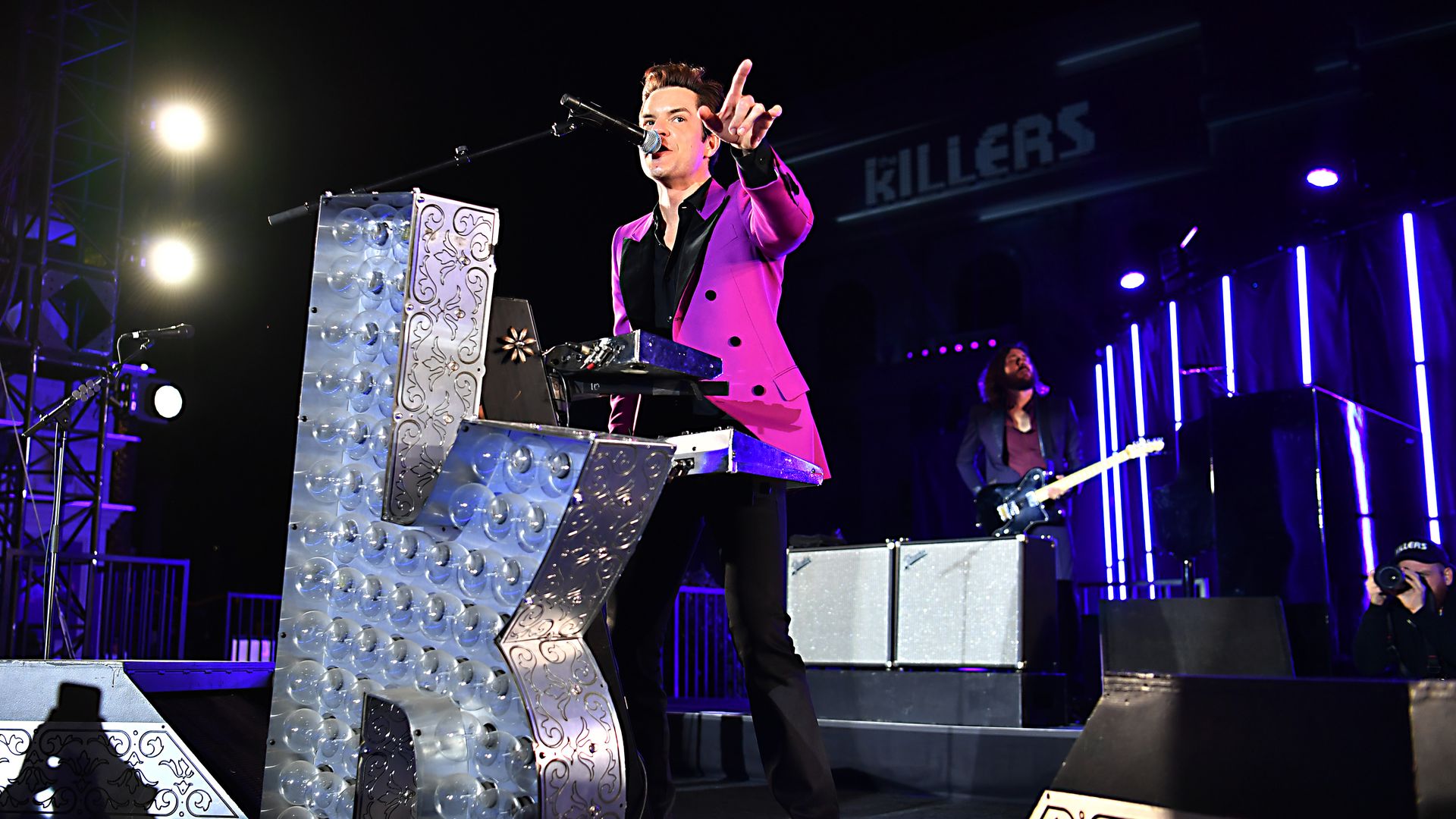 Brandon Flowers of The Killers plays keyboard and points while singing into a microphone in front of a large light-up "K"