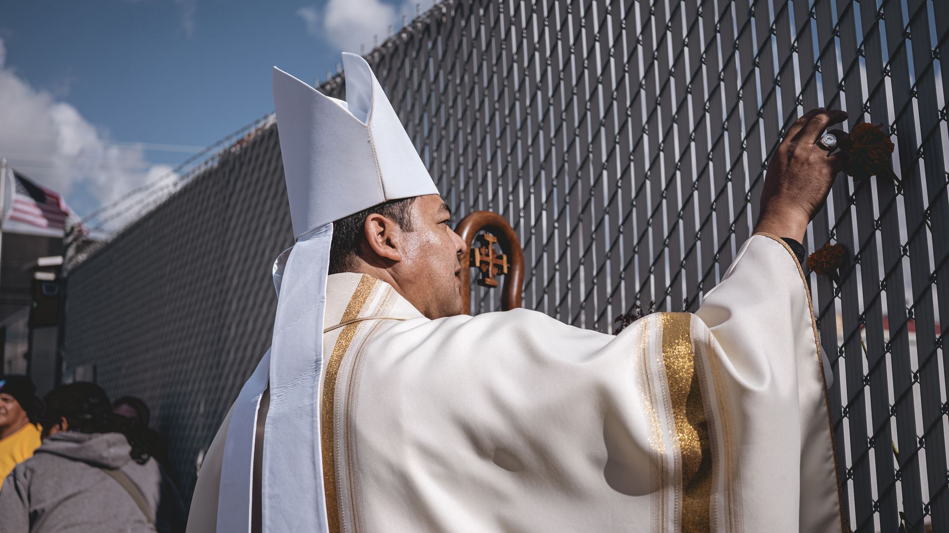A man in white religious robes with gold trim and a tall white hat places orange flowers on a metal fence under a blue sky with clouds, holding a wooden staff with a cross.