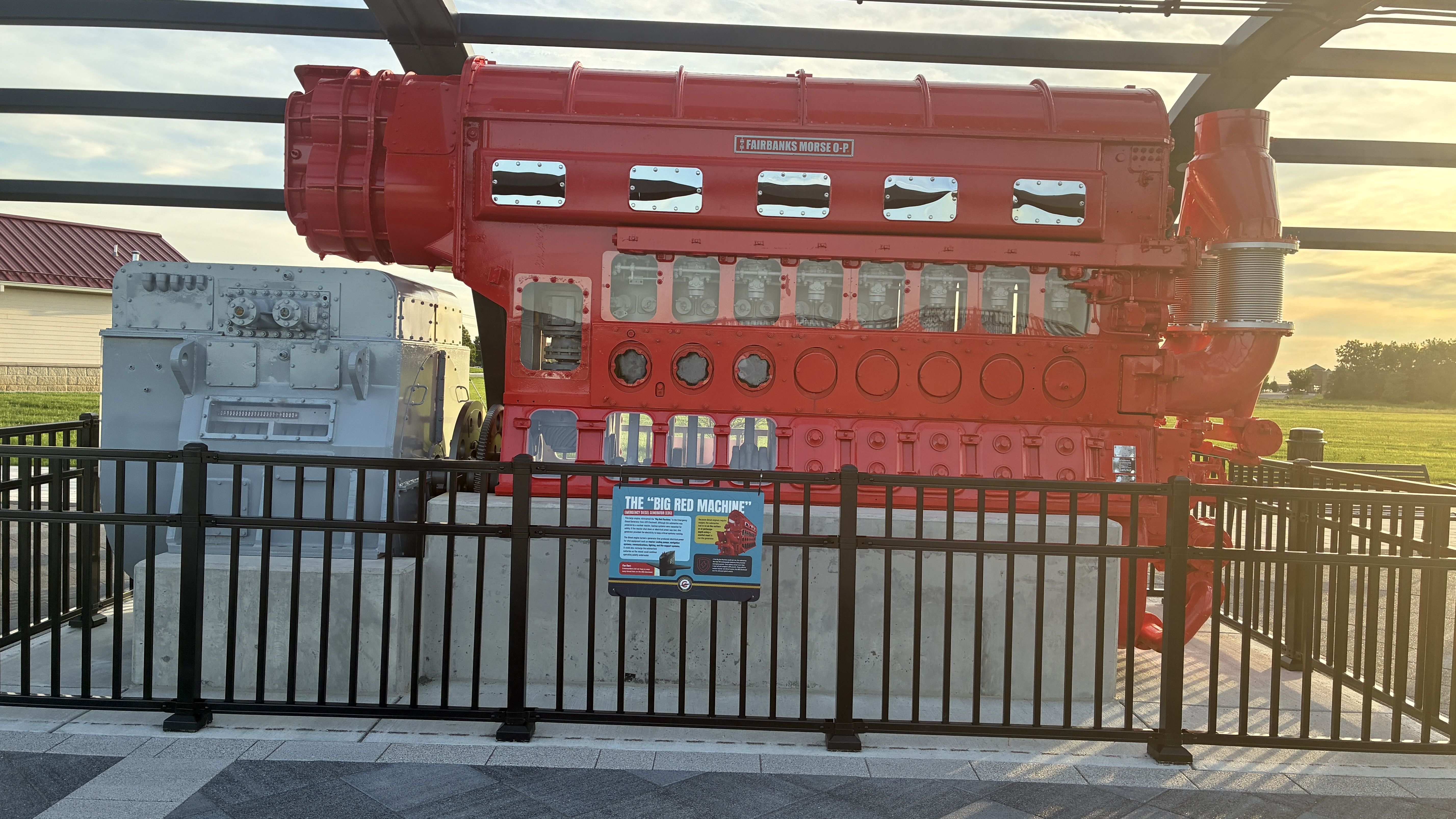 Large red Fairbanks Morse O-P engine on display behind a black fence, with a gray-blue auxiliary unit on the left and a field at sunset in the background; sign reads THE BIG RED MACHINE.