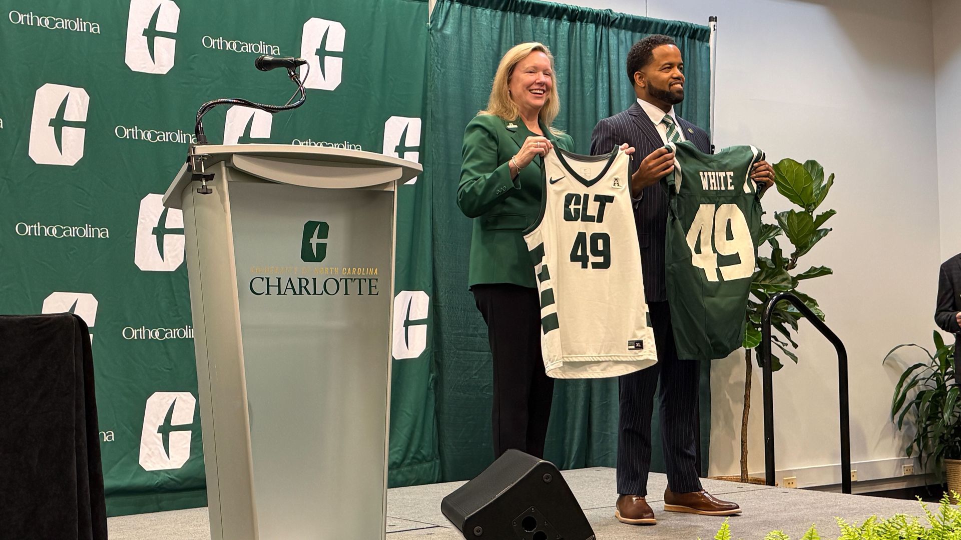 Two people hold Charlotte 49ers jerseys on stage with green backdrop and podium labeled University of North Carolina Charlotte during an event.