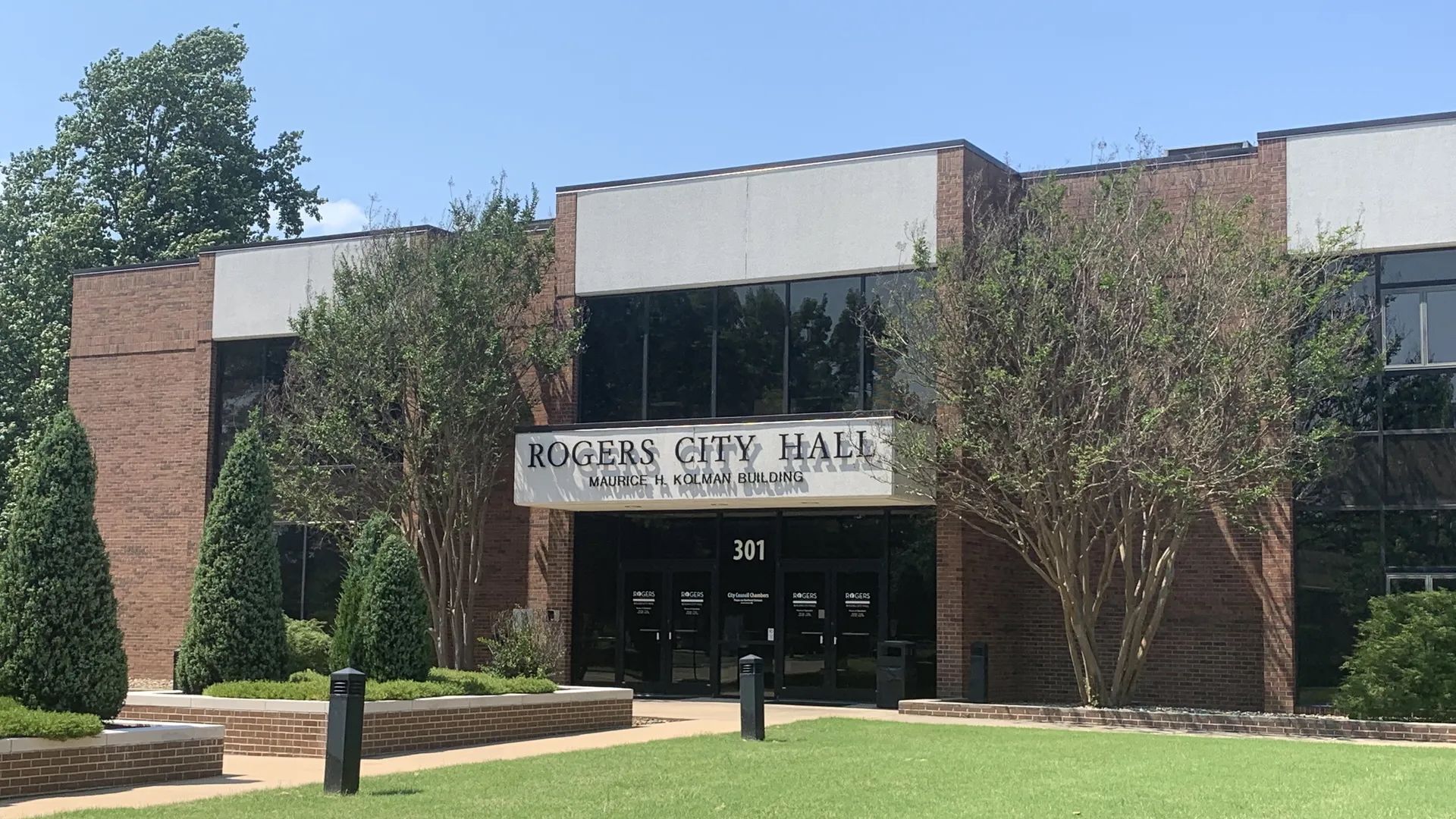 Brick municipal building labeled "Rogers City Hall" with glass entrance doors, trees, and a lawn in front.