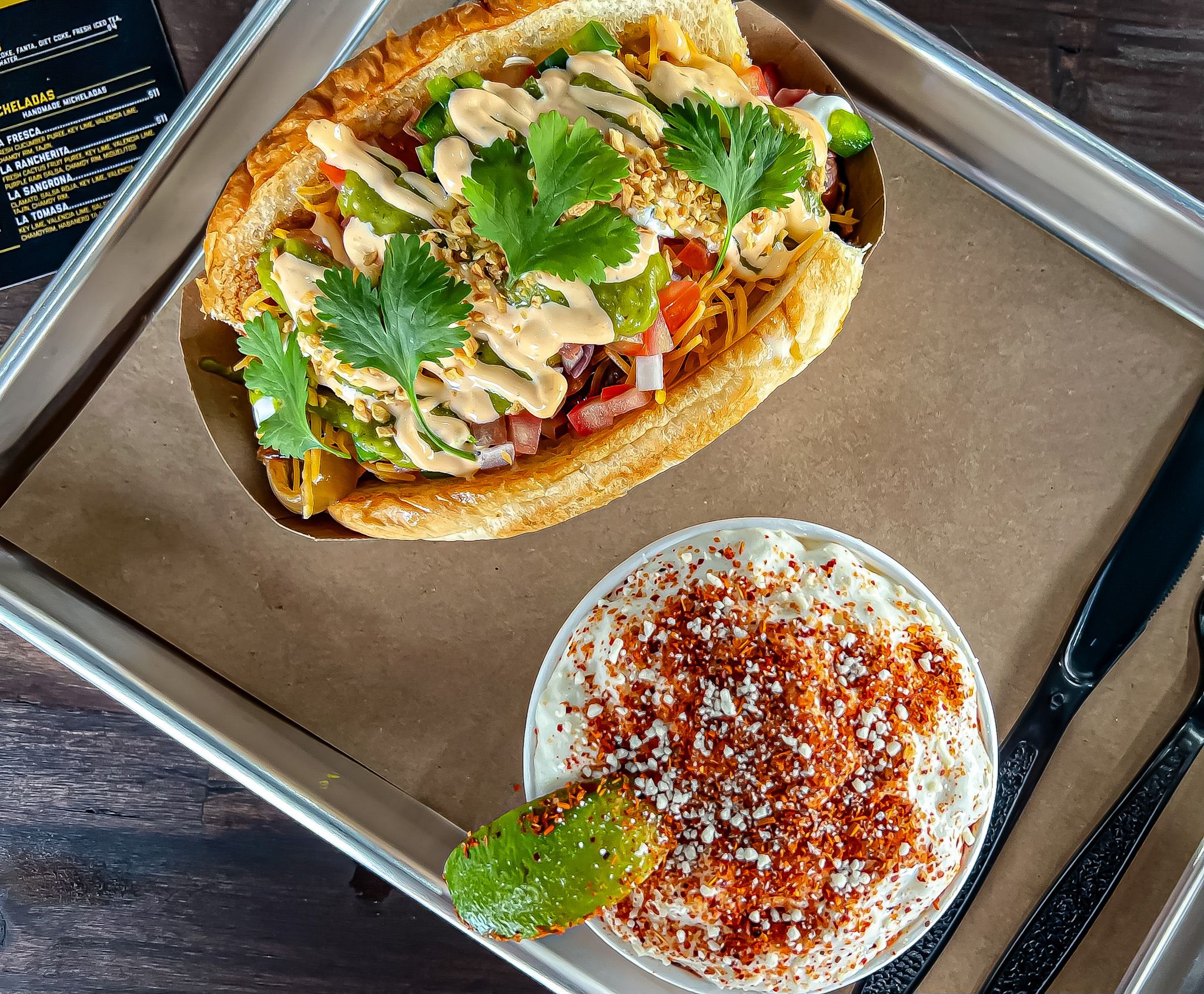 A view from above of a hot dog covered in aioli and cilantro and a side of elote in a bowl.