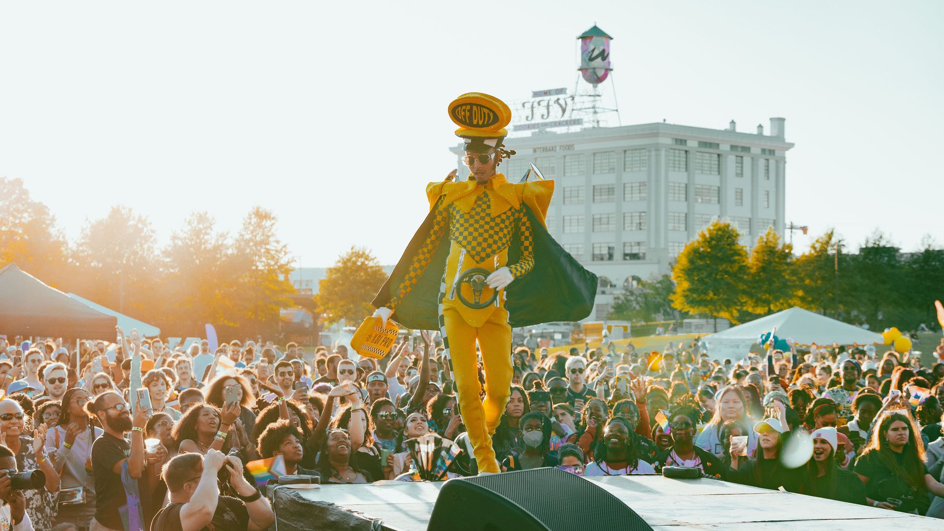 Person in yellow and black checkered costume with large hat and cape stands on stage holding a steering wheel, facing a large crowd outdoors in daylight with a white building and trees in background.