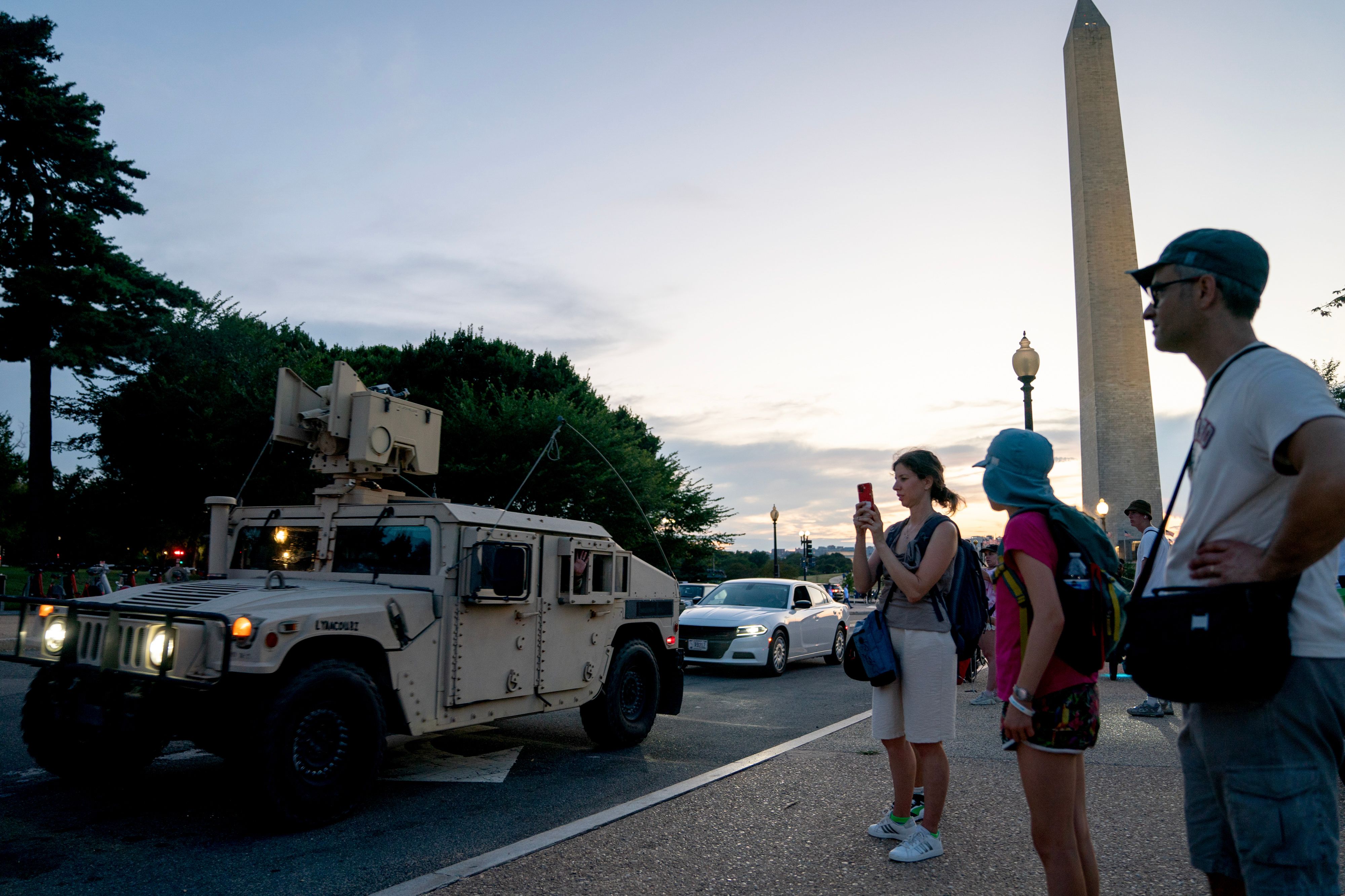 People standing near a military Humvee on a street at dusk with the Washington Monument in the background.
